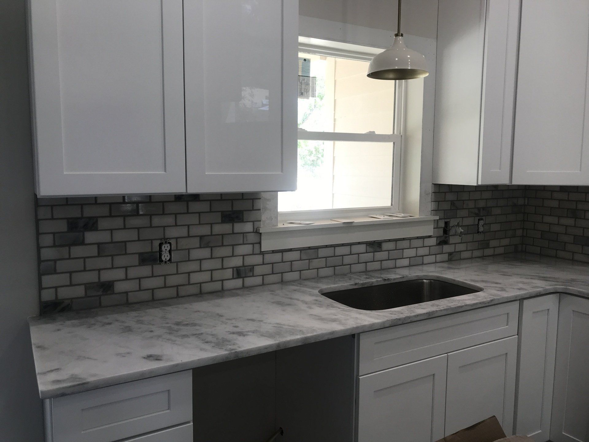White kitchen with marble countertops, gray backsplash, and a window above the sink.