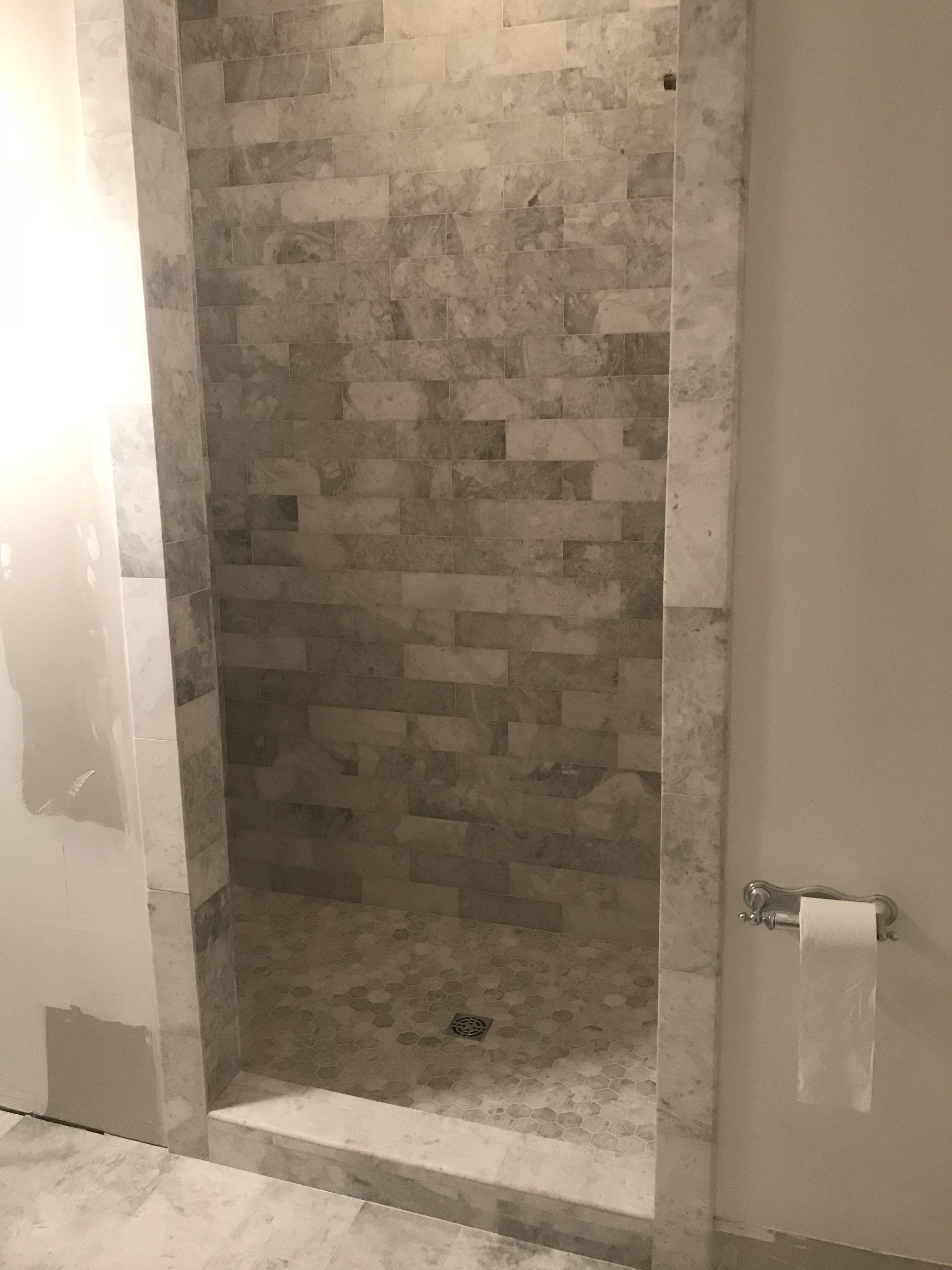 Shower stall with gray brick tile and a white marble frame. A roll of toilet paper is visible.