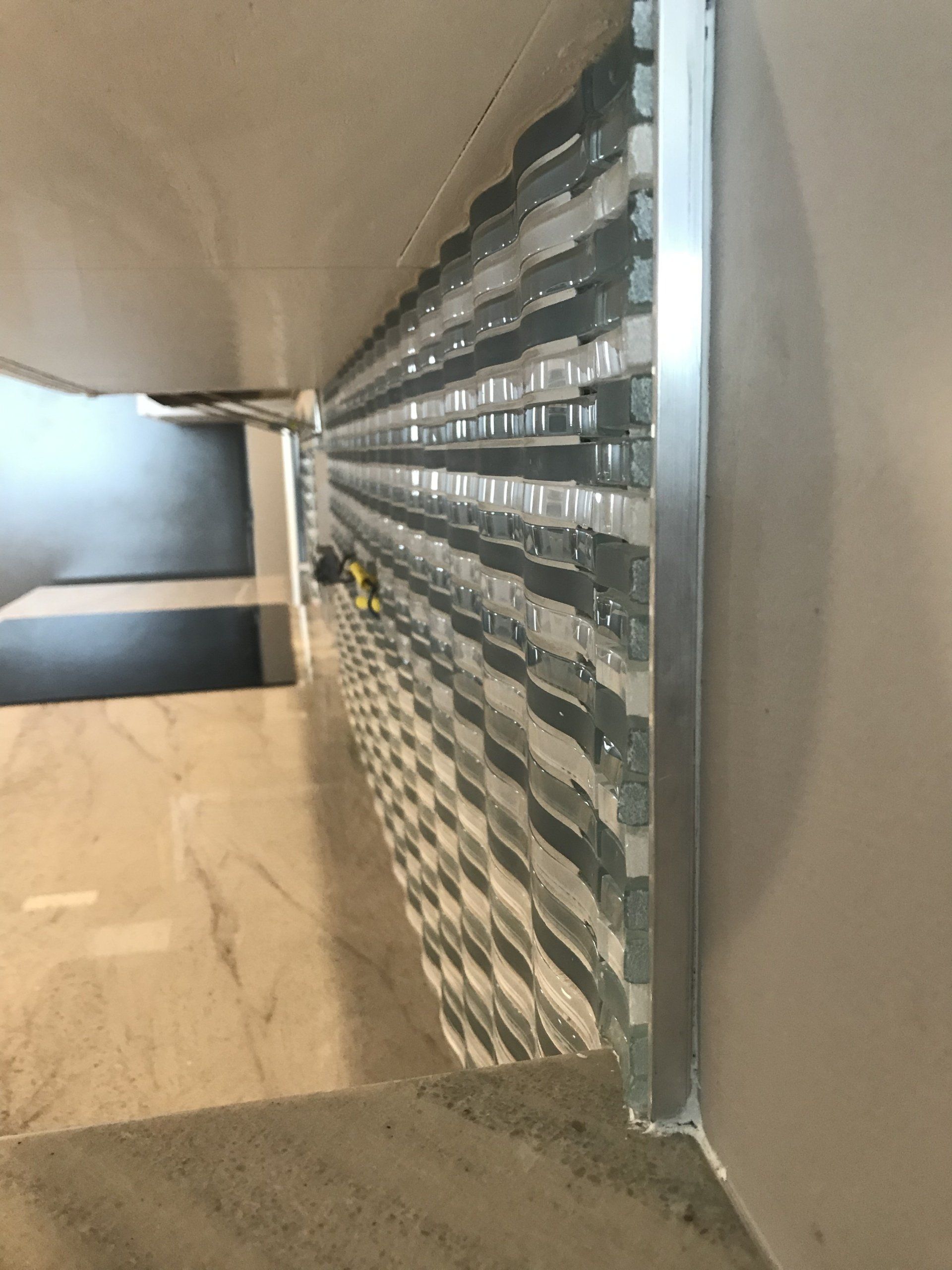 Close-up of a tile backsplash with a metal edge. Light gray, white, and blue tiles are installed below a countertop.