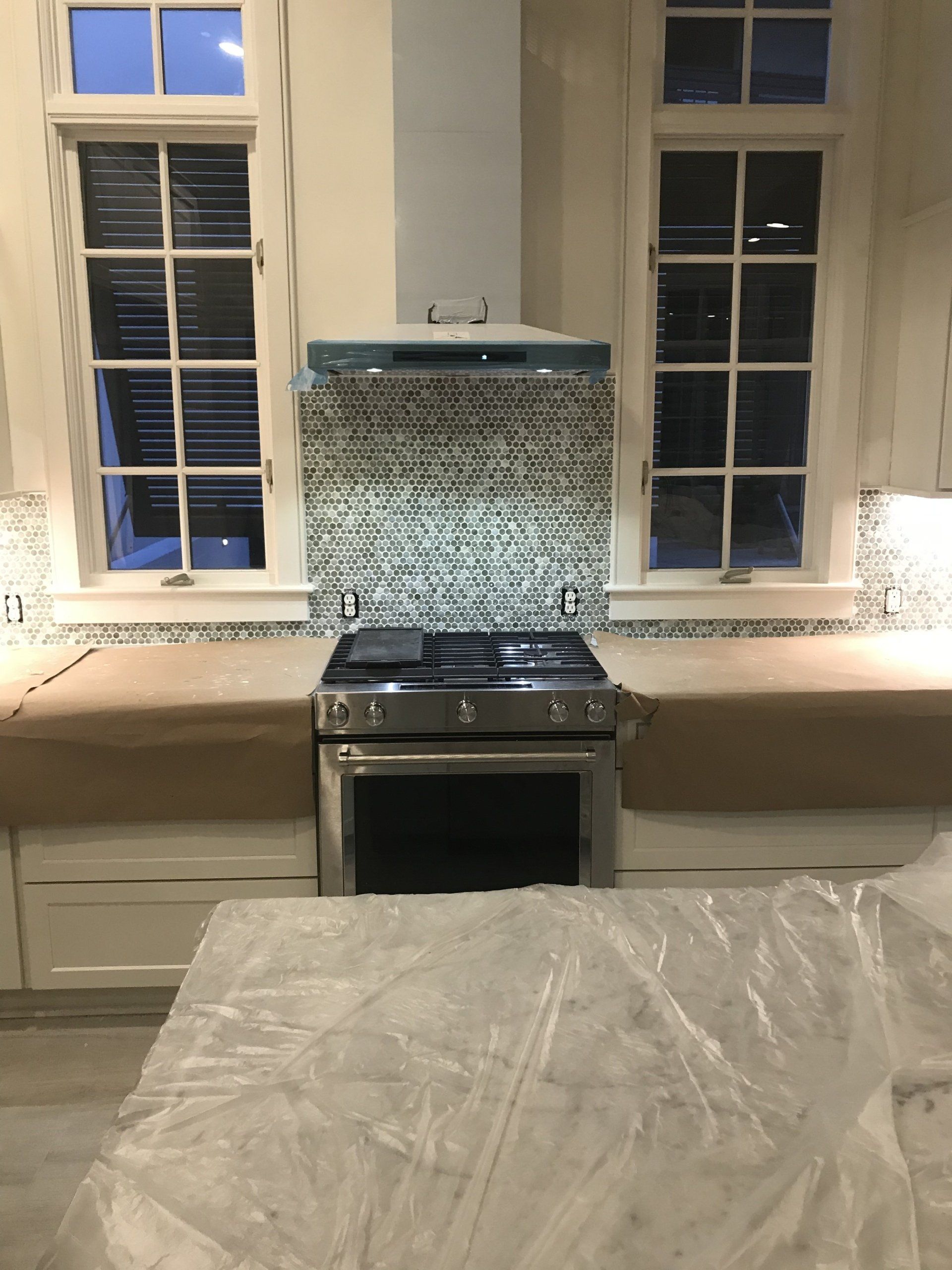 Kitchen with stove, backsplash, and hood, flanked by windows. Countertops and island covered in protective plastic.