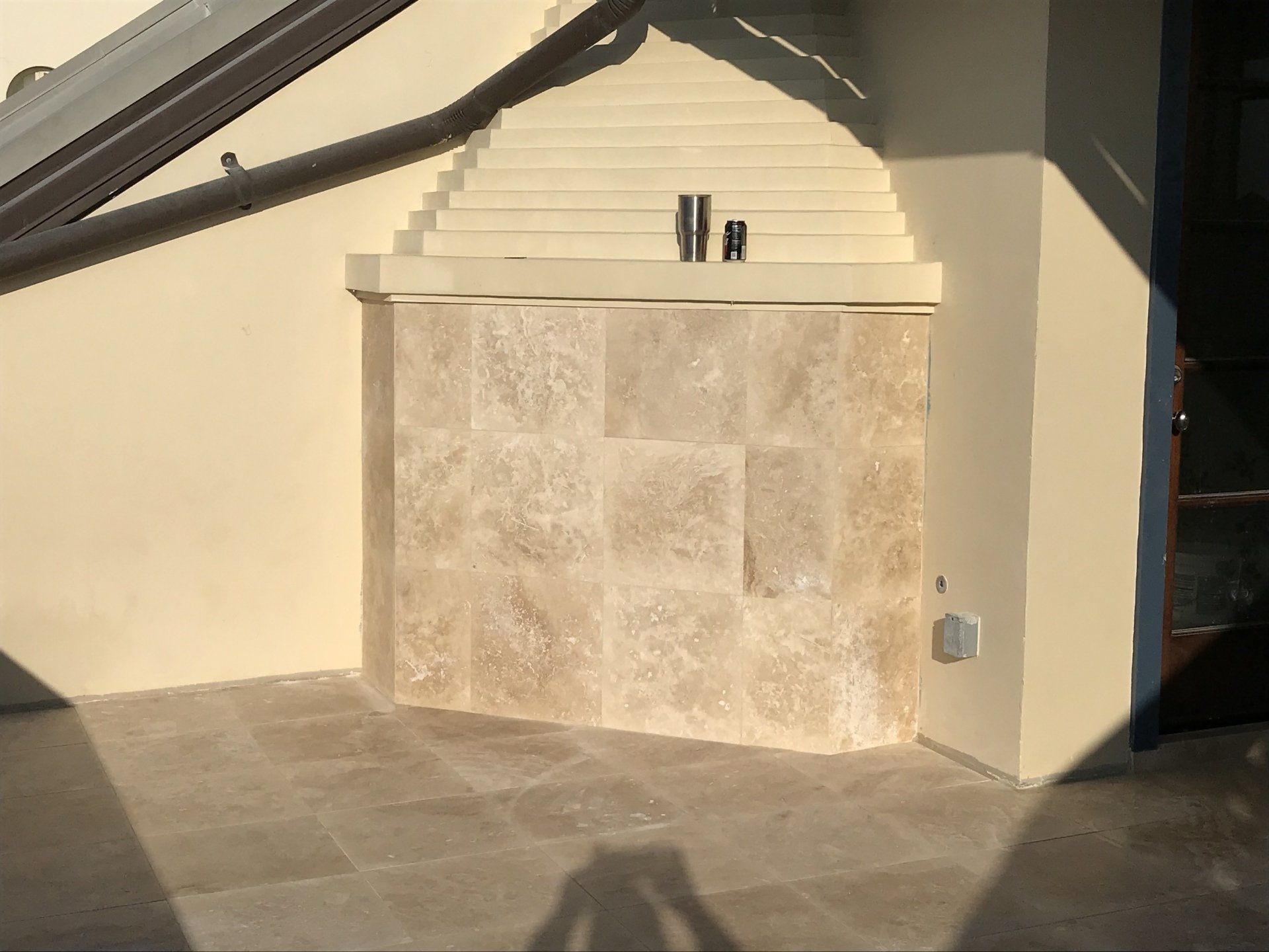 Light-colored stone wall enclosure on patio with two metal pipes, neutral paint, sunny outdoor setting.