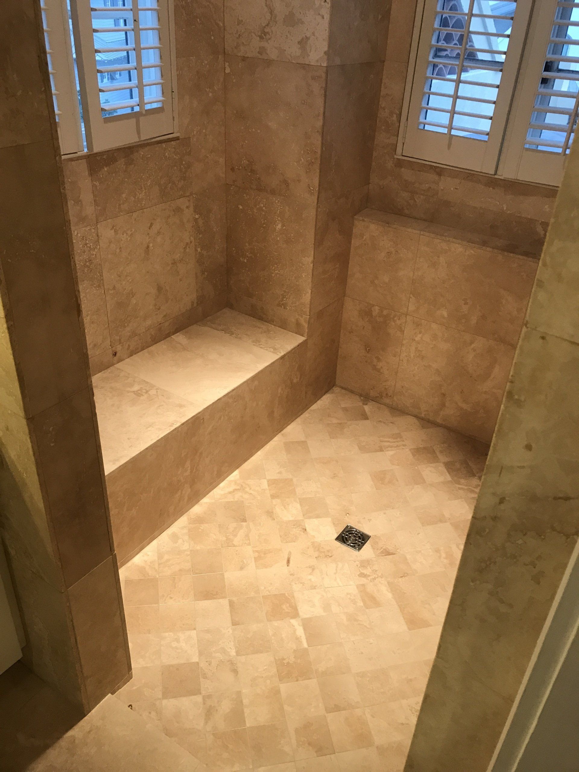 A tiled shower stall with a built-in bench. Beige tiles, two windows.