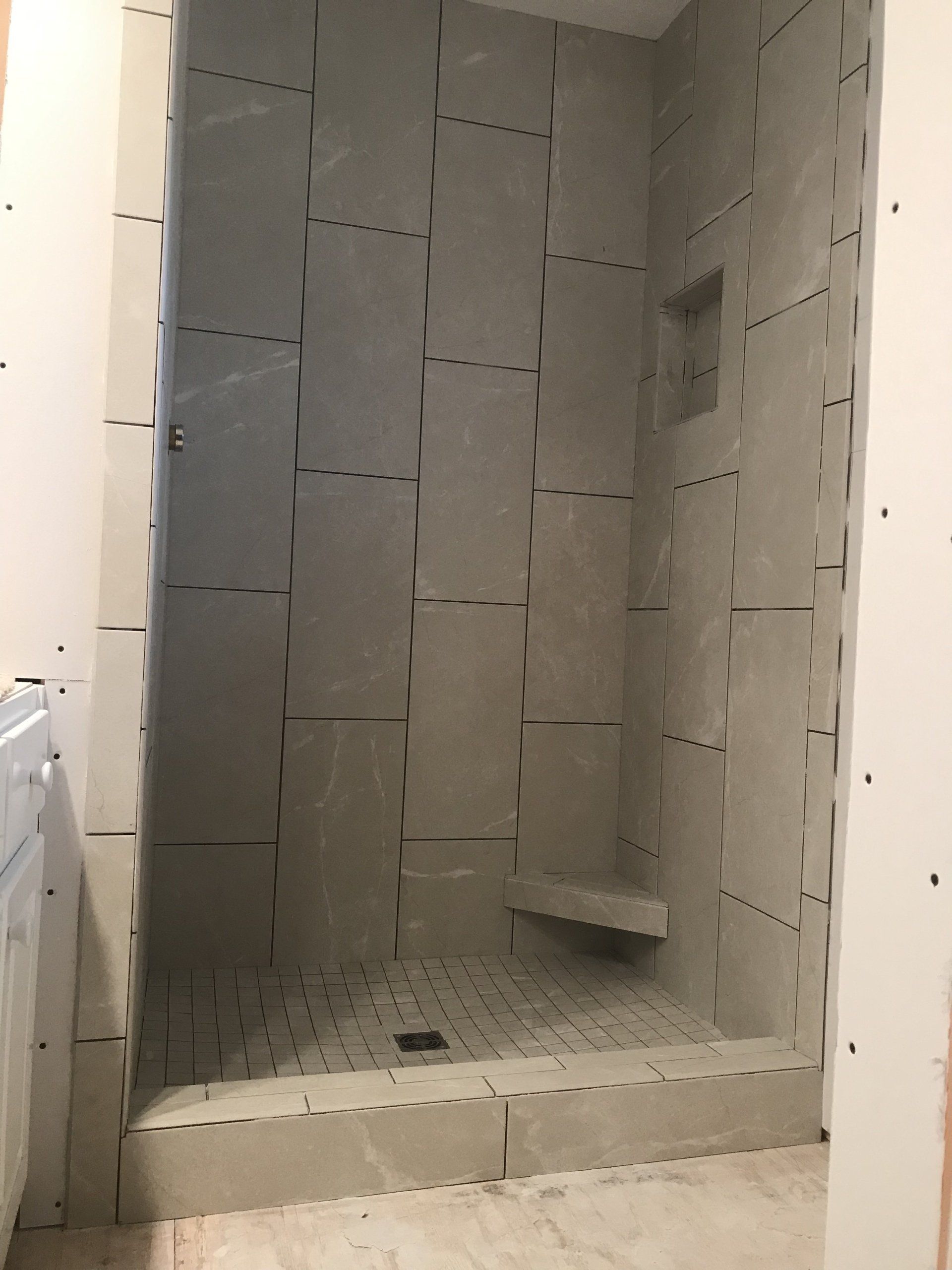 Shower stall under construction, gray walls with tile, built-in bench, and floor drain.