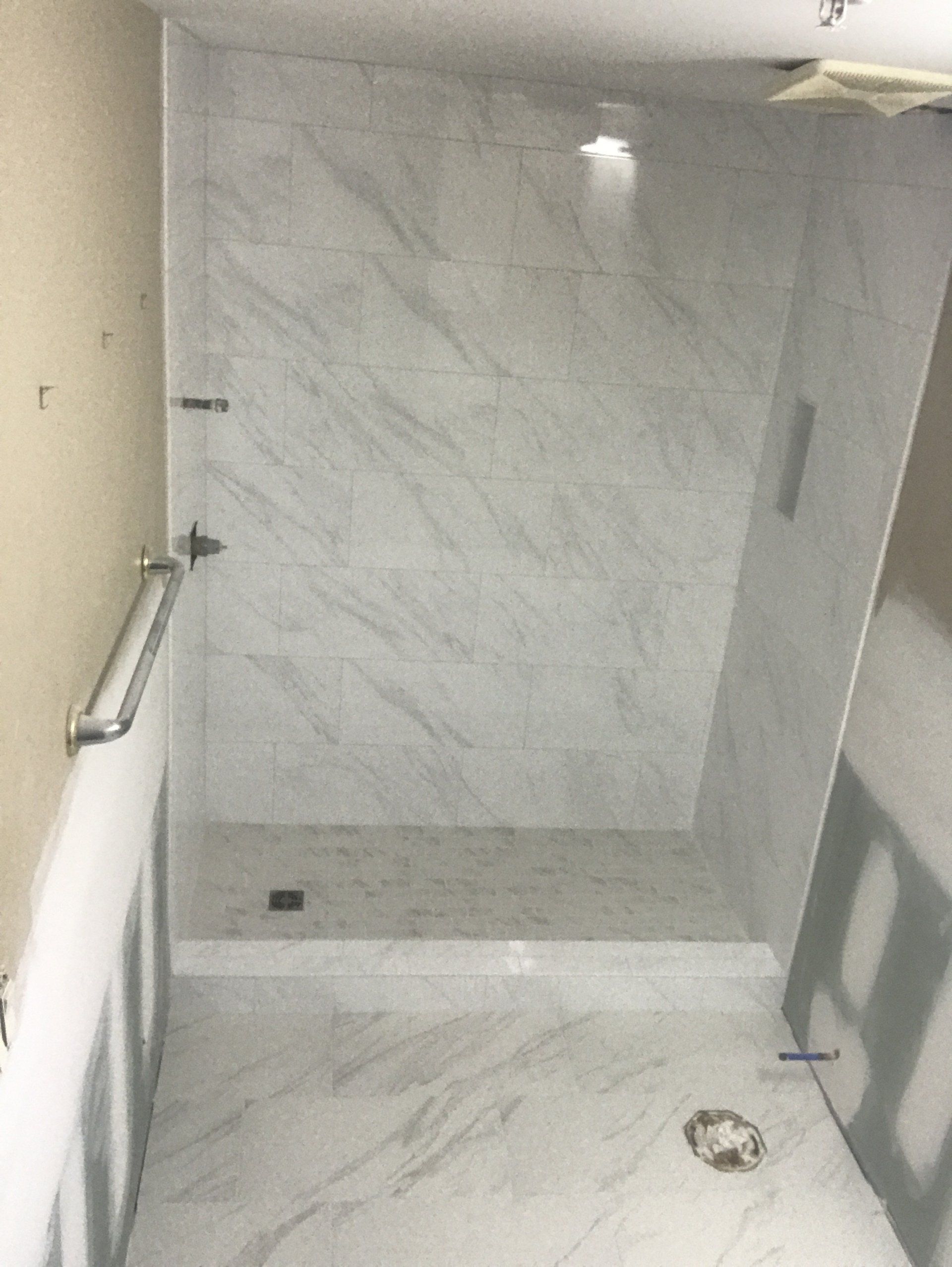 Shower stall with marble-look tile, glass door, grab bar, and drain, under construction.