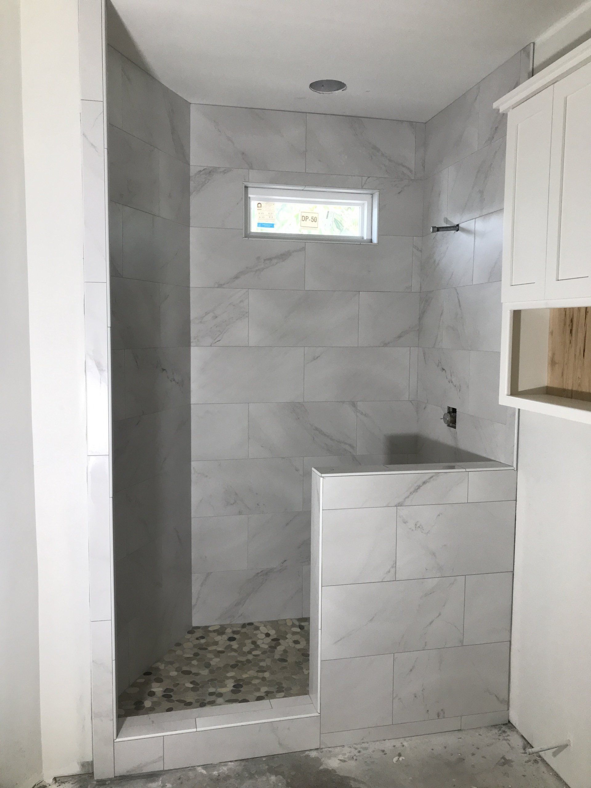 A walk-in shower with gray marble-look tile, pebble floor, small window, and shower head, in a bathroom.