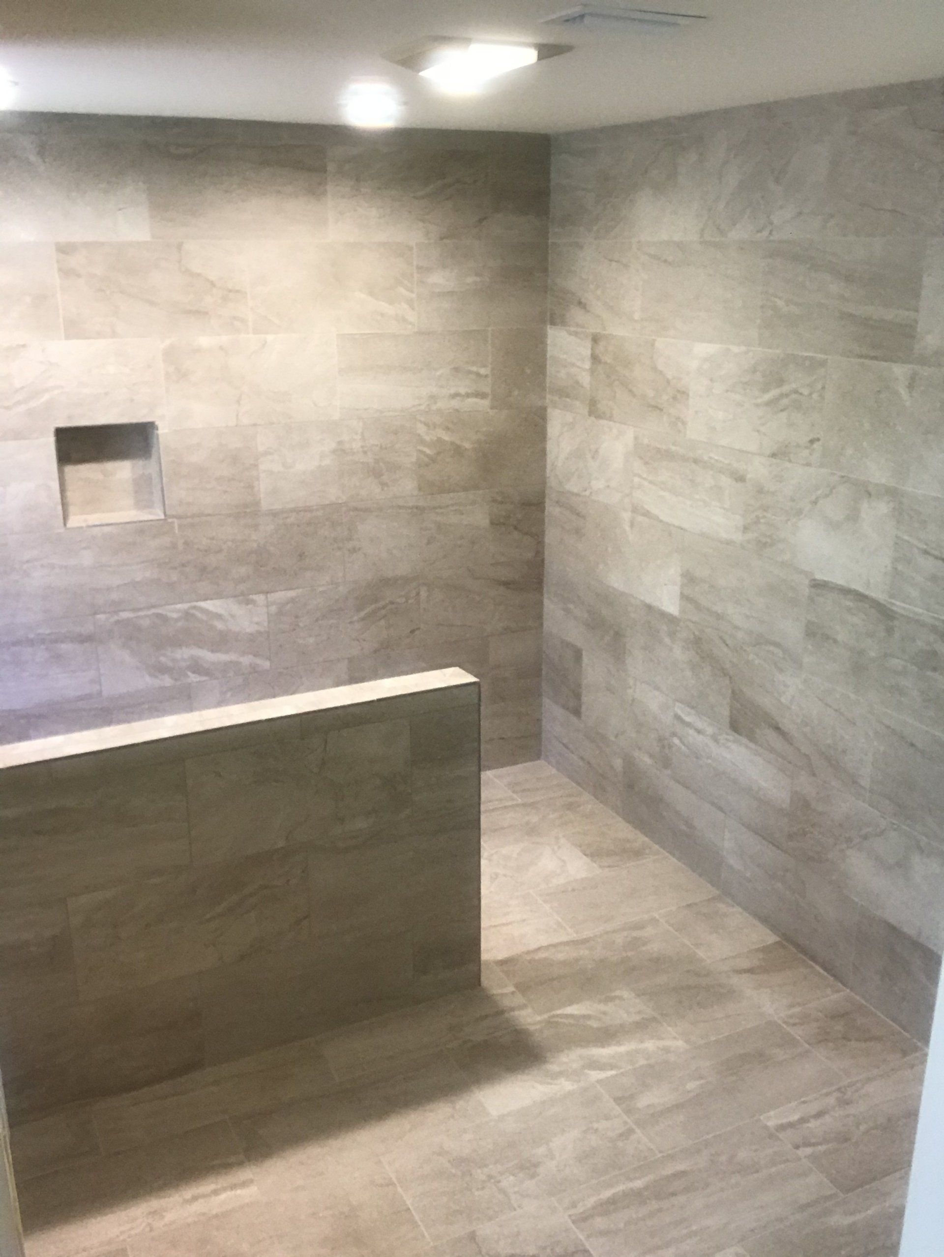 Gray tiled shower with a low wall, recessed shelf, and bright overhead lights.