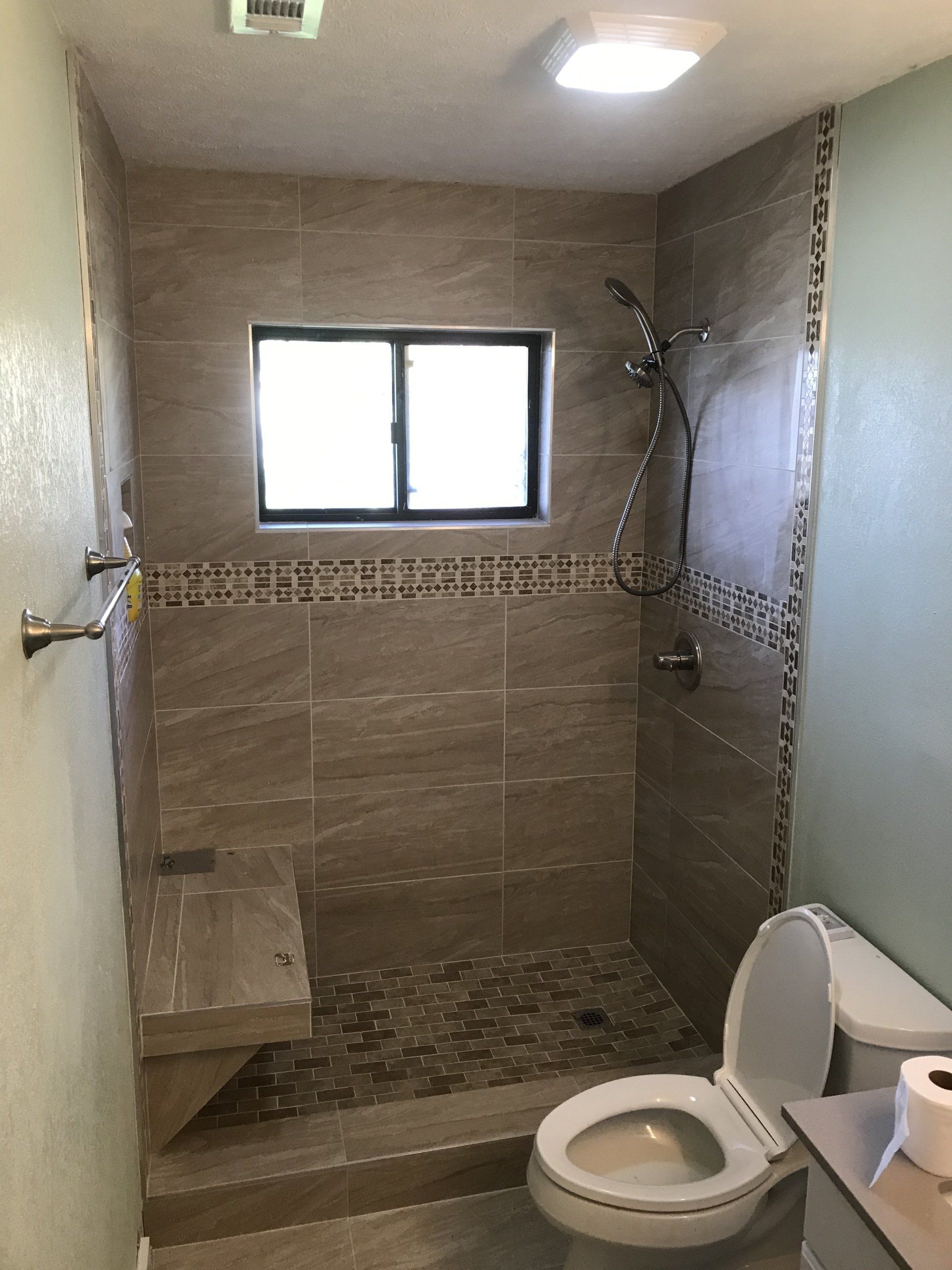 Bathroom with tiled shower, toilet, and bench seat. Neutral tones, small window, and pebble floor.