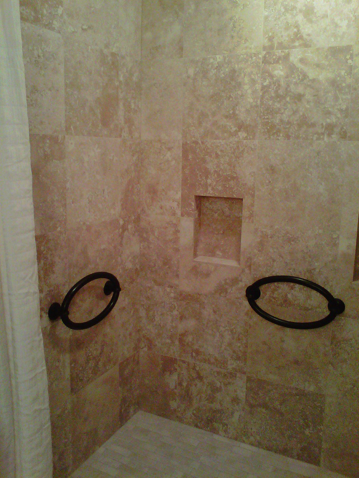 Bathroom shower with tan tile walls, black towel rings, and a recessed shelf.