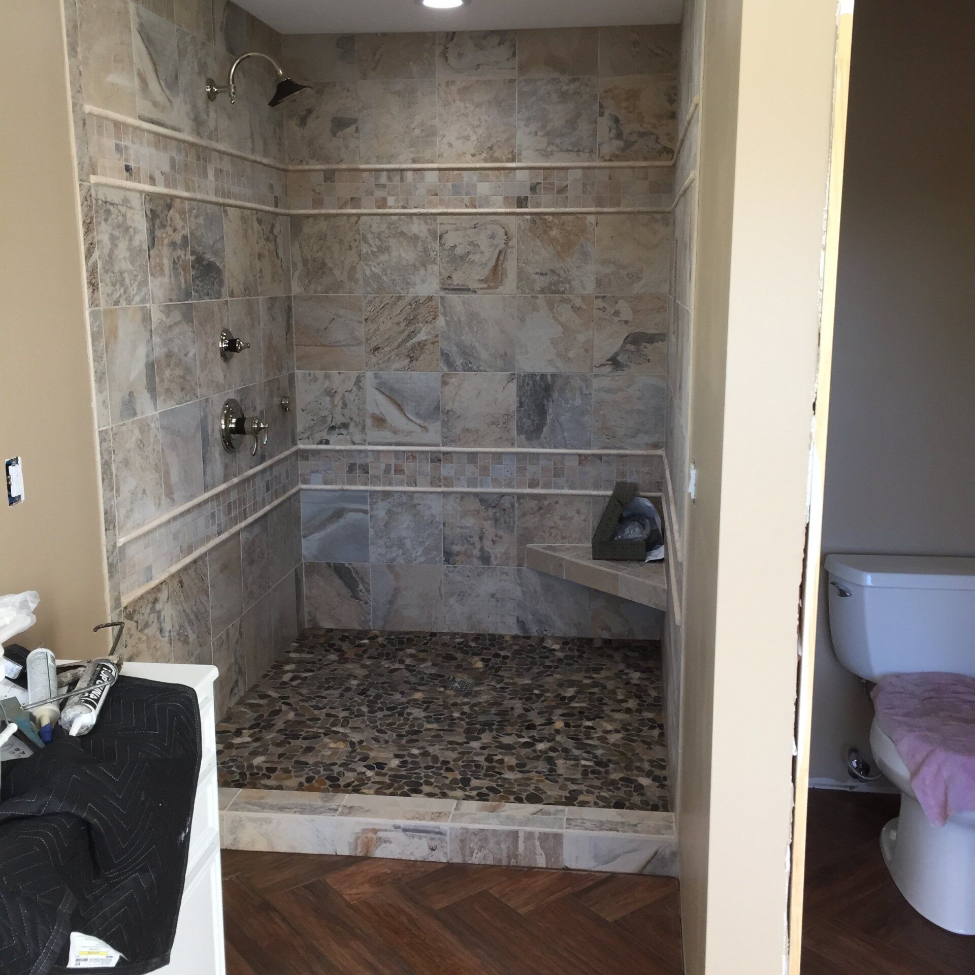 Newly tiled shower with pebble floor, built-in seat, and bronze fixtures, next to a toilet.