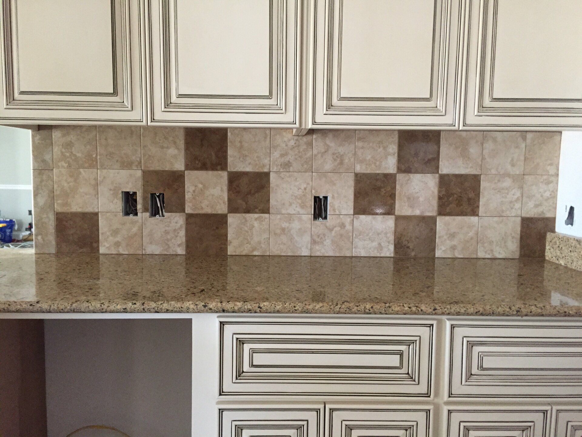 Kitchen backsplash with alternating light and dark brown square tiles, light brown countertop, and cream cabinets.