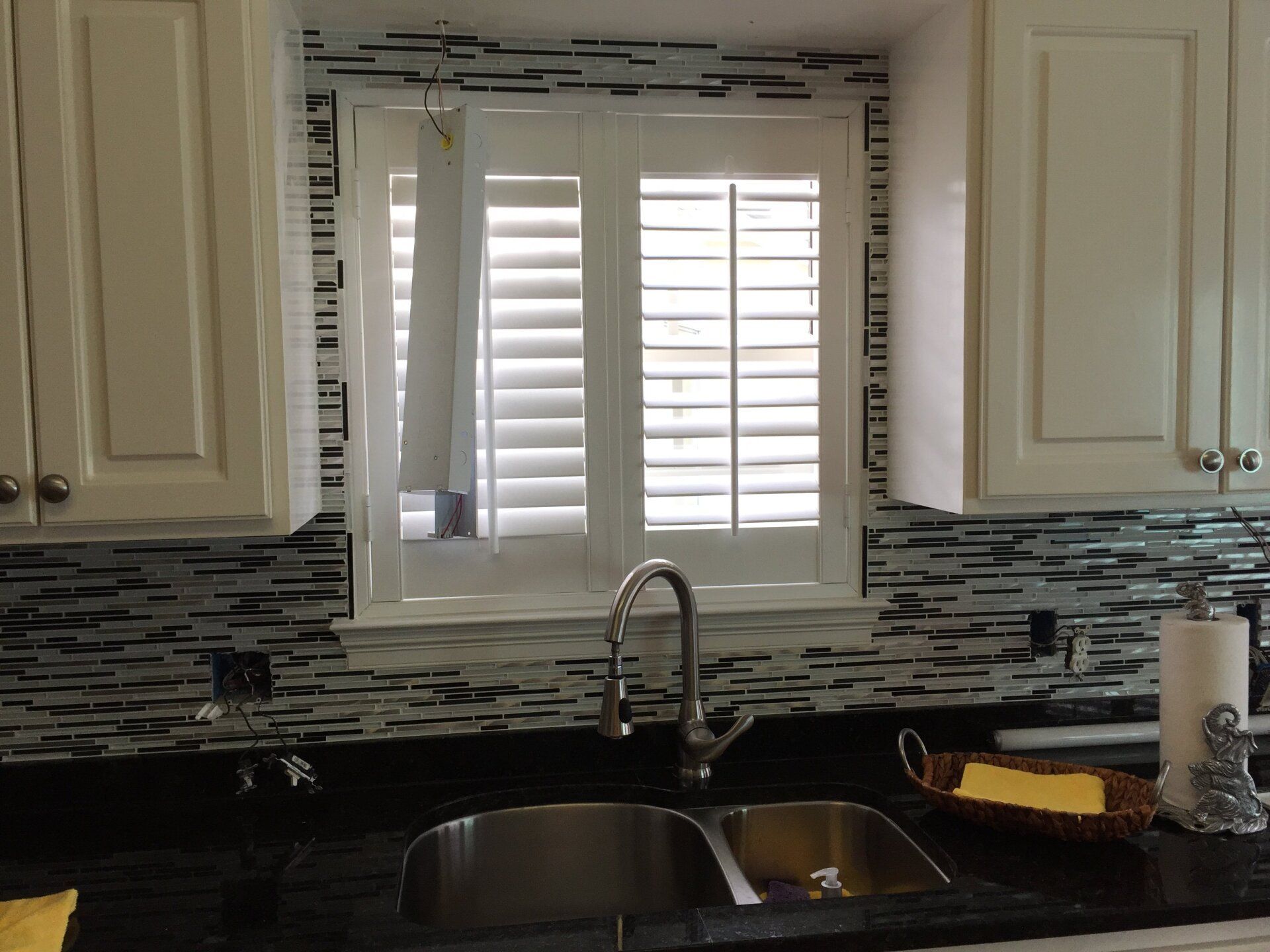Kitchen sink under a window with white shutters, black countertops, and a mosaic backsplash.
