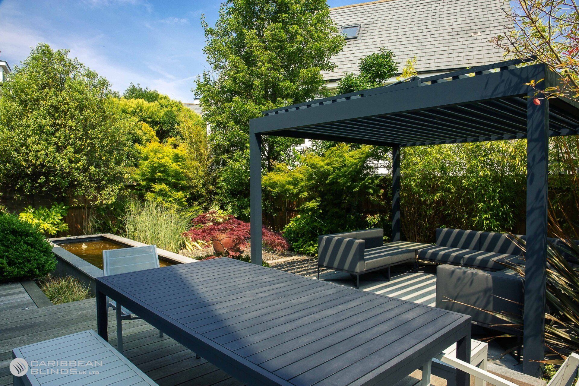 Modern outdoor patio with a dark pergola, matching dining table and sofa set, a small reflecting pool, and lush greenery.