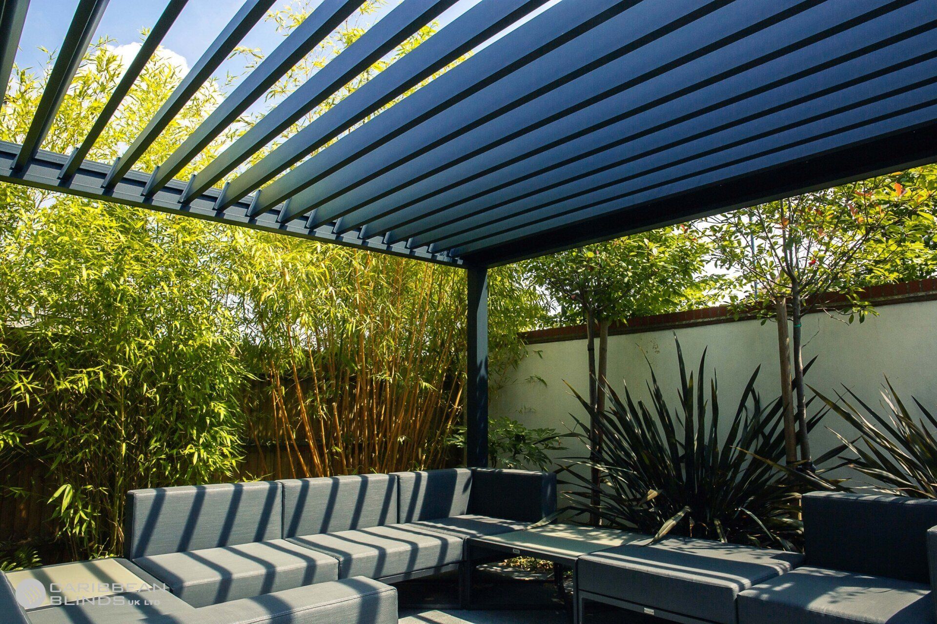A dark grey outdoor sectional sofa under a modern louvered pergola, situated in a garden with bamboo and dark foliage.