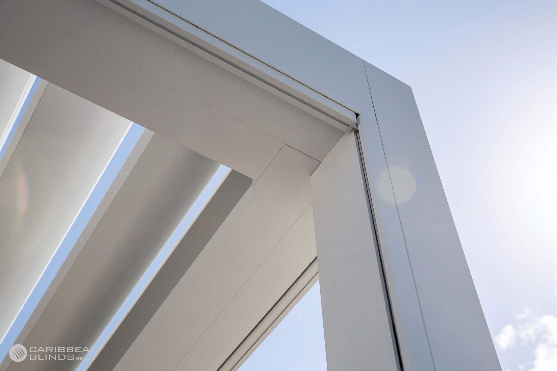 A low-angle view of a white, modern pergola structure with an adjustable louvered roof against a clear, sunny sky.