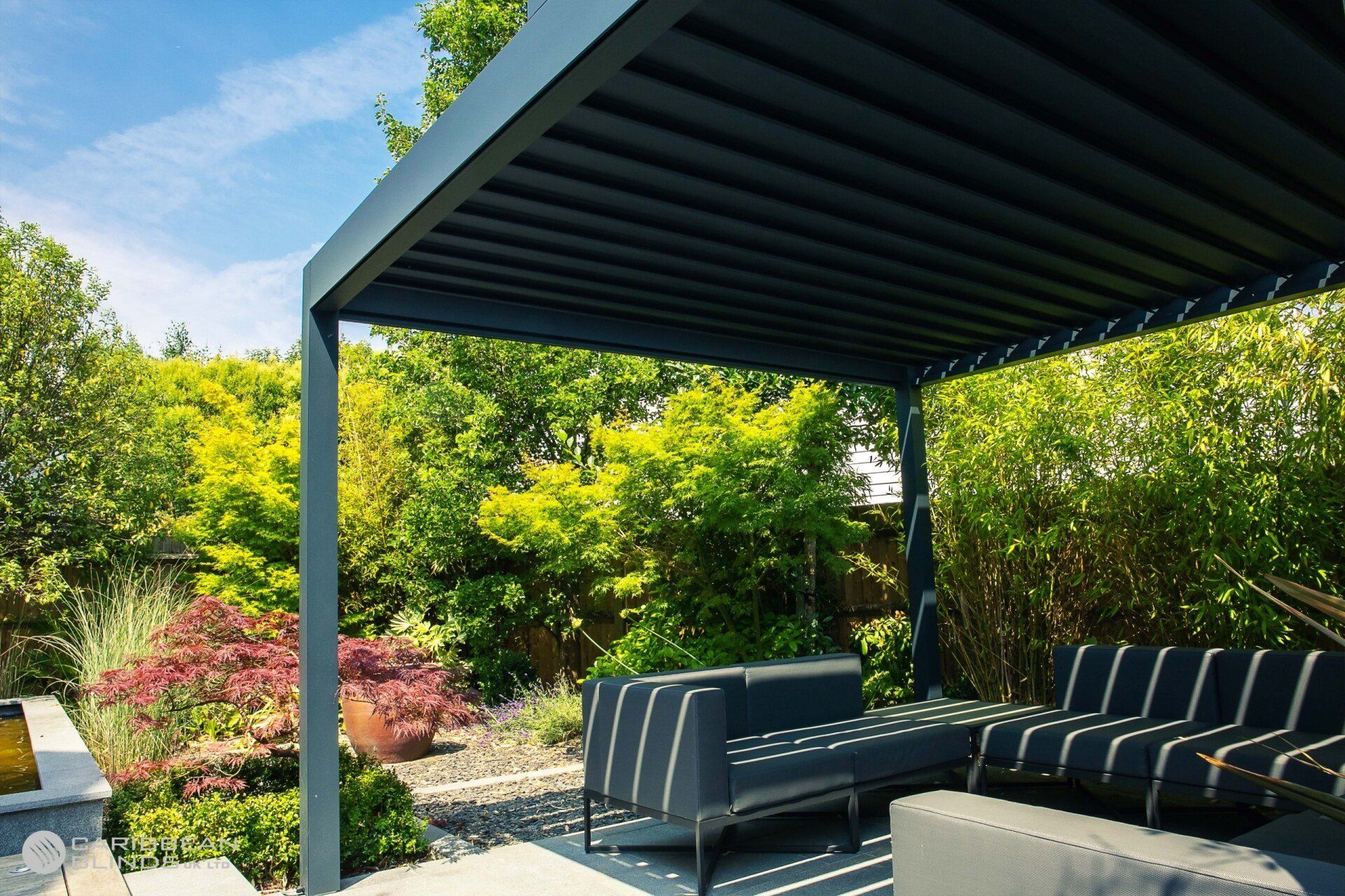 A modern, grey louvered pergola shades a contemporary sofa set on a patio, surrounded by lush green garden foliage.