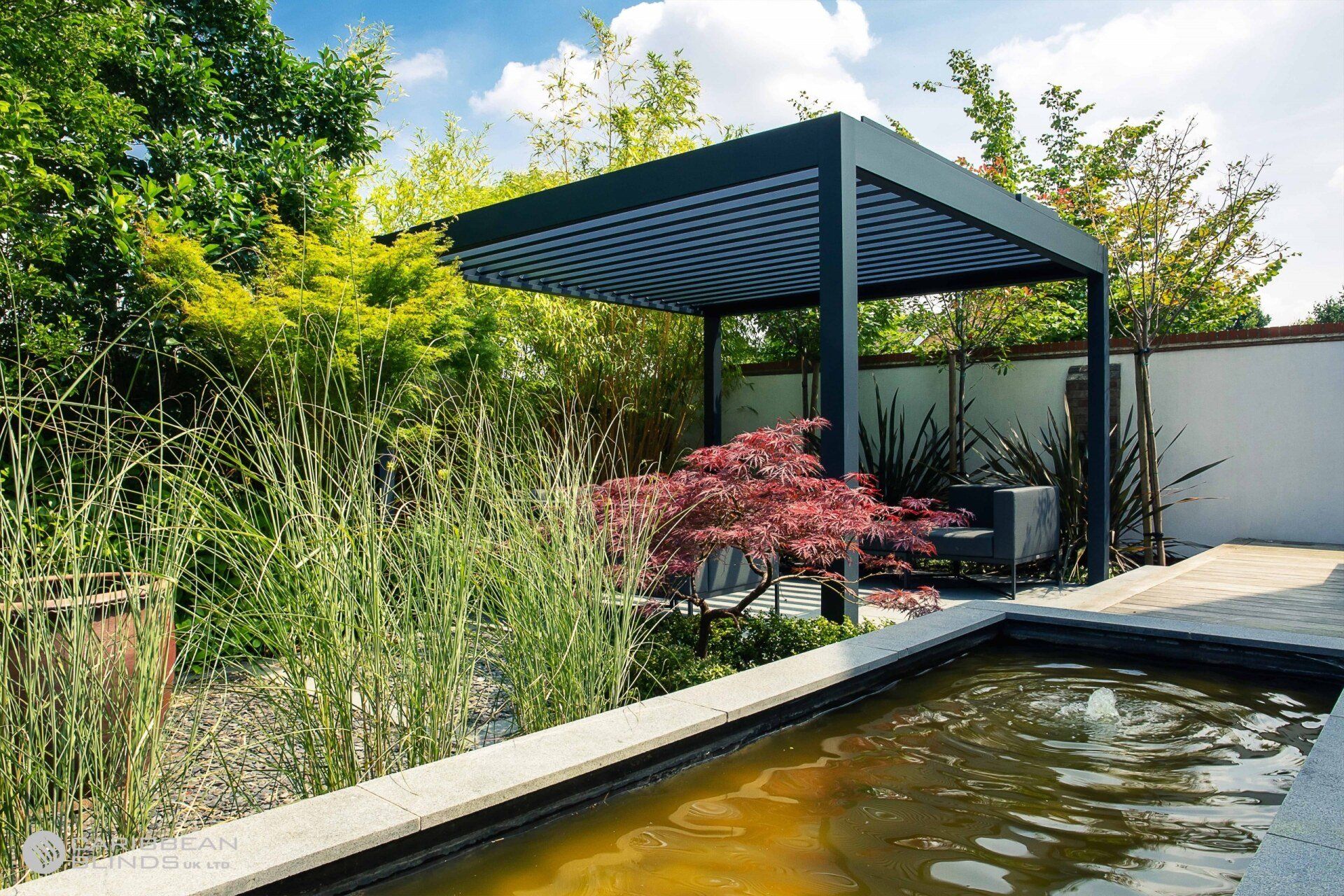 A dark gray pergola stands in a garden beside a reflecting pool, surrounded by lush green foliage and a red maple tree.