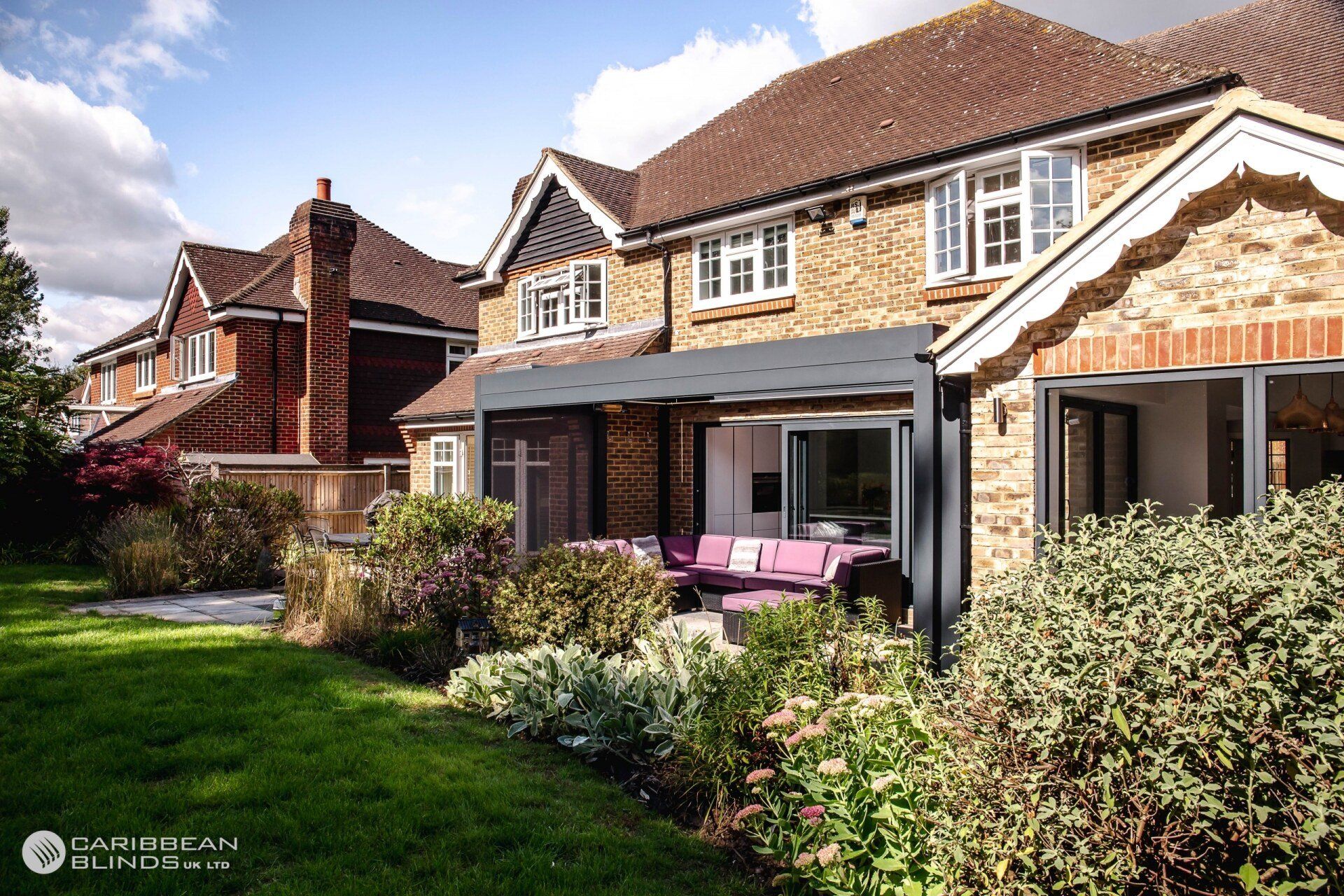 A modern dark gray pergola extends over a patio with purple seating attached to a brick house on a sunny day.