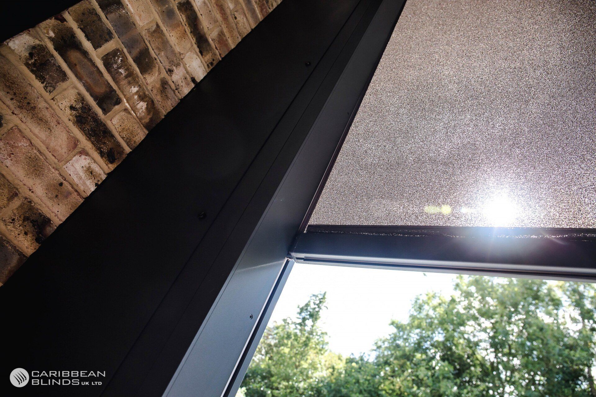 A close-up of a black window frame installed next to an exposed brick wall, with a partial view of a gray solar shade.