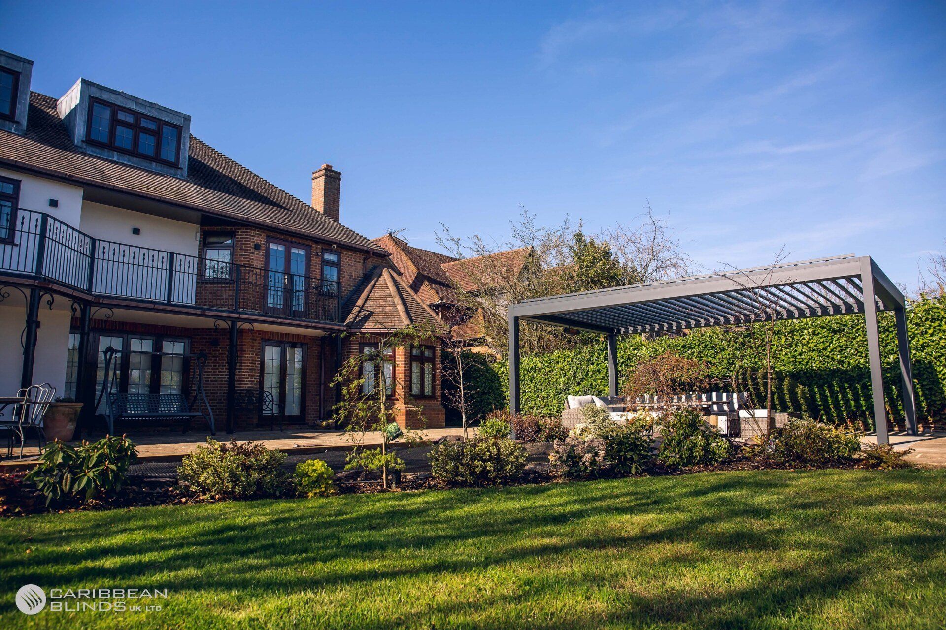 A brick house with a balcony and a backyard patio featuring a modern metal pergola over seating under a sunny blue sky.