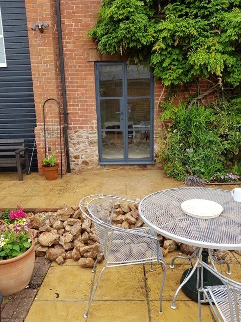 A brick wall with a grey door and a nearby pile of rocks on a patio, featuring a metal bistro table and potted plants.