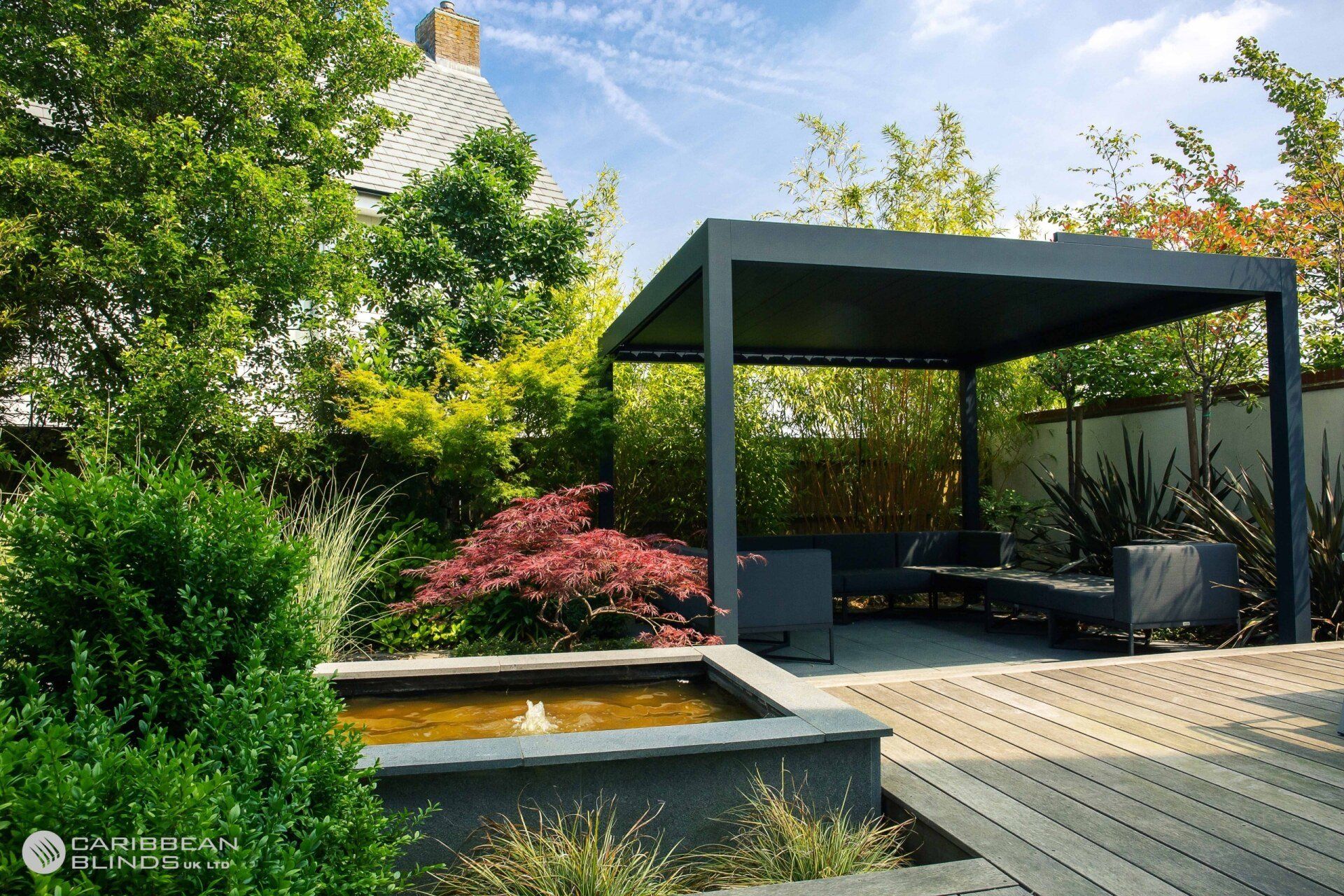 A modern garden featuring a dark pergola over lounge furniture, a stone water feature, and lush green plants.