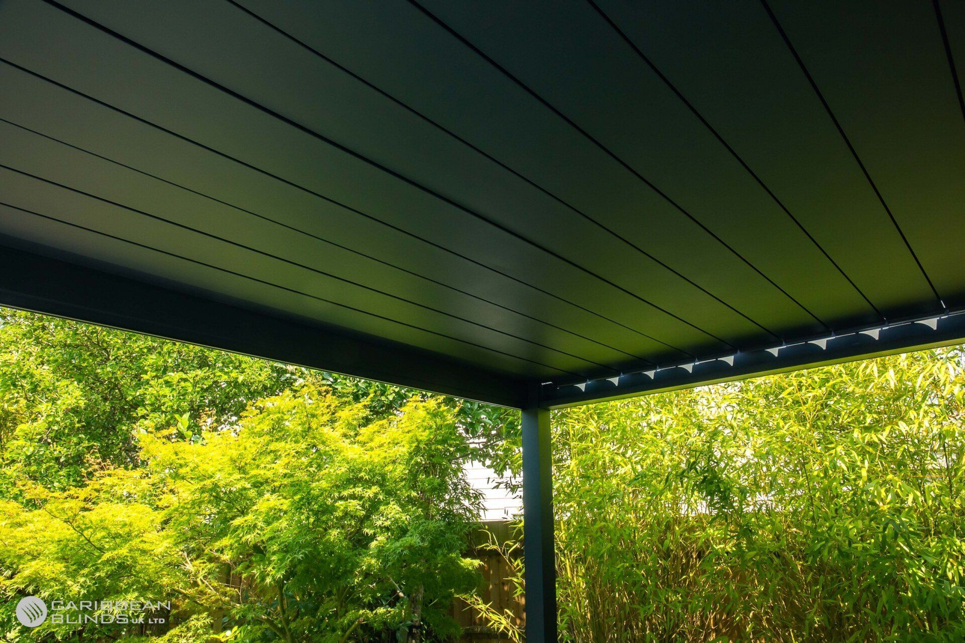 A dark-colored, modern louvered patio roof structure surrounded by lush green foliage.