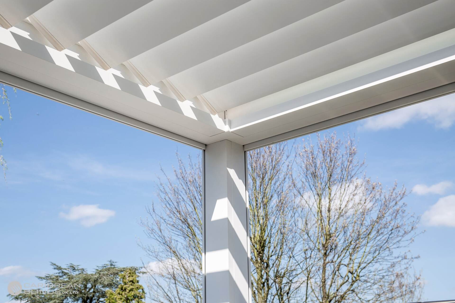 A white louvered patio roof structure set against a blue sky, with sunlight creating geometric shadows on the support post.