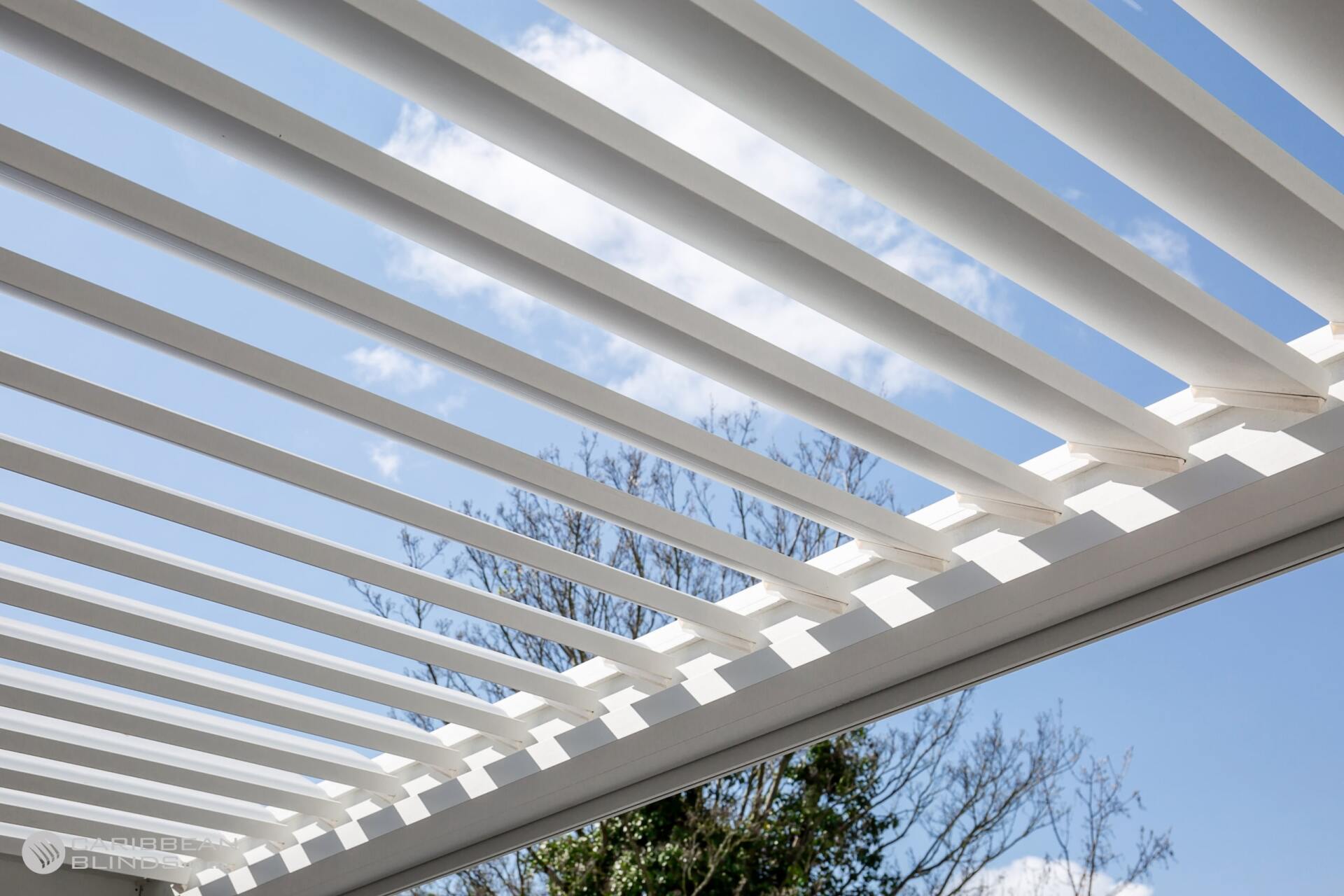 Low-angle view of a modern white louvered pergola roof with open slats against a blue sky with clouds and trees.