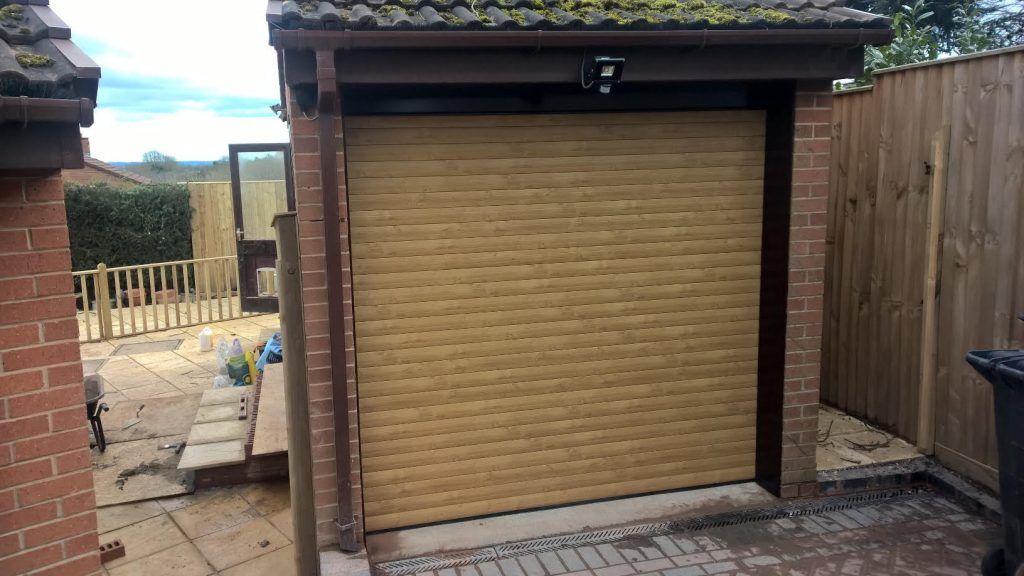 A tan-colored, horizontally ribbed garage door set in a brick exterior building with a wooden fence to the side.