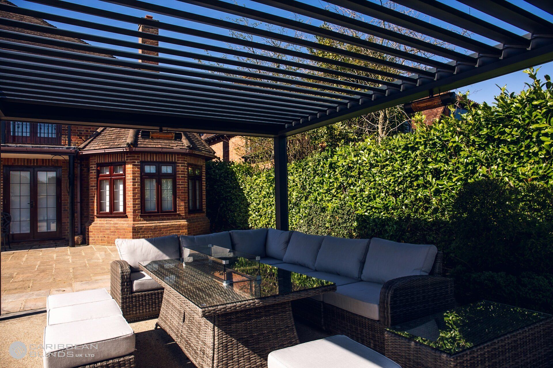 A wicker sectional sofa and glass-topped table sit under a modern black louvered pergola next to a brick house and hedge.
