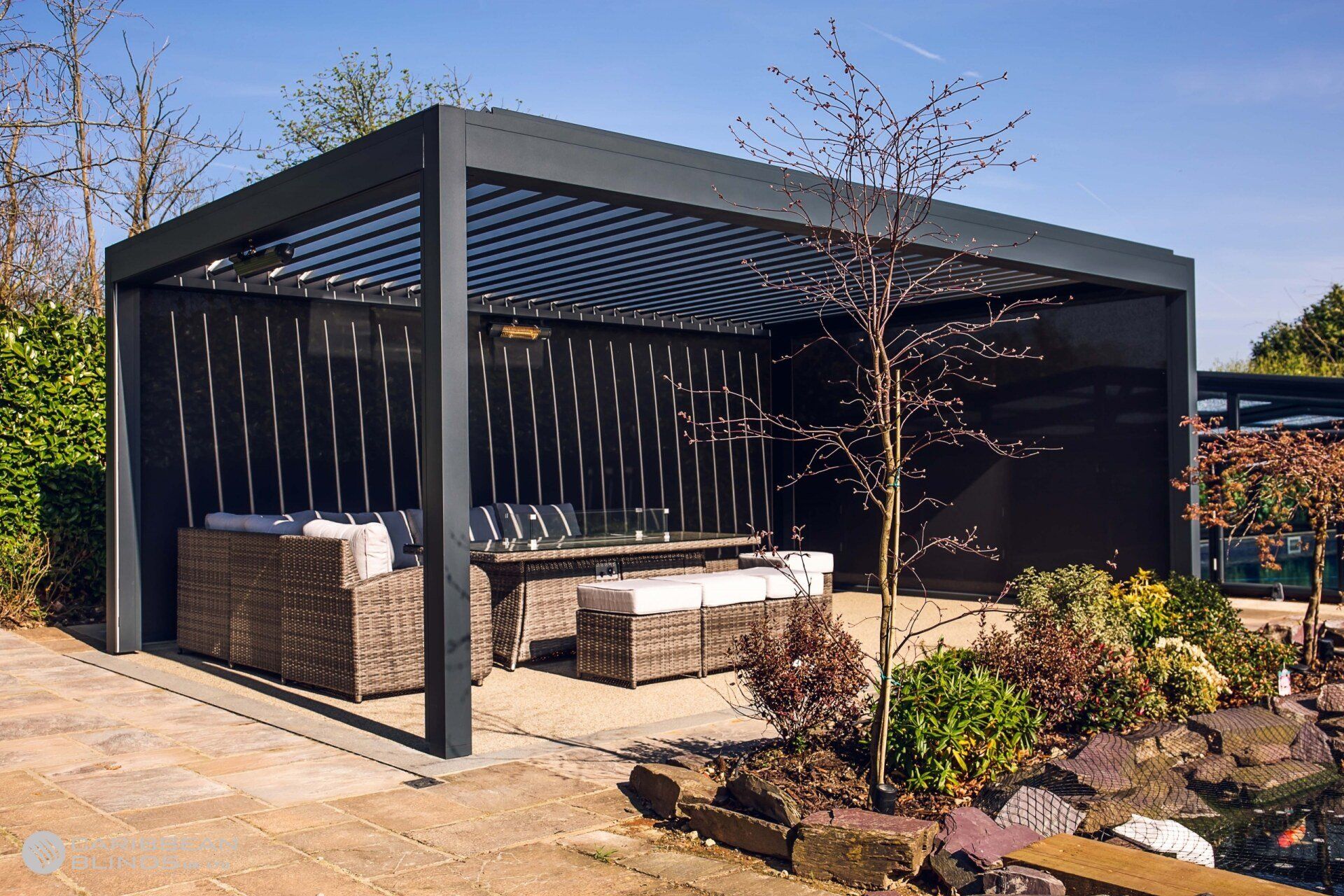 A modern black louvered pergola structure outdoors, featuring comfortable wicker lounge seating on a stone patio.