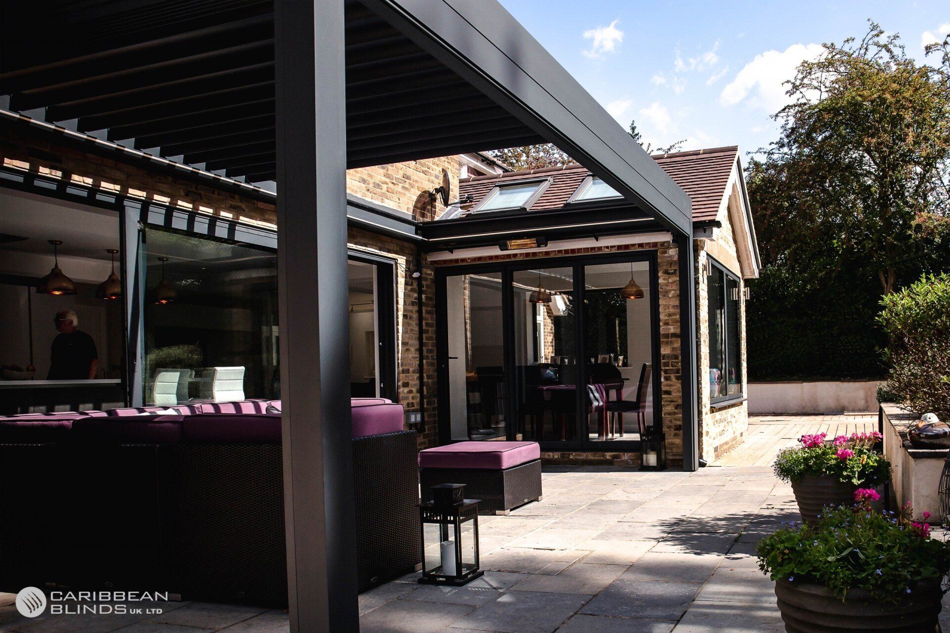 A dark, louvered patio pergola extends over a stone terrace with outdoor seating and potted plants next to a house.