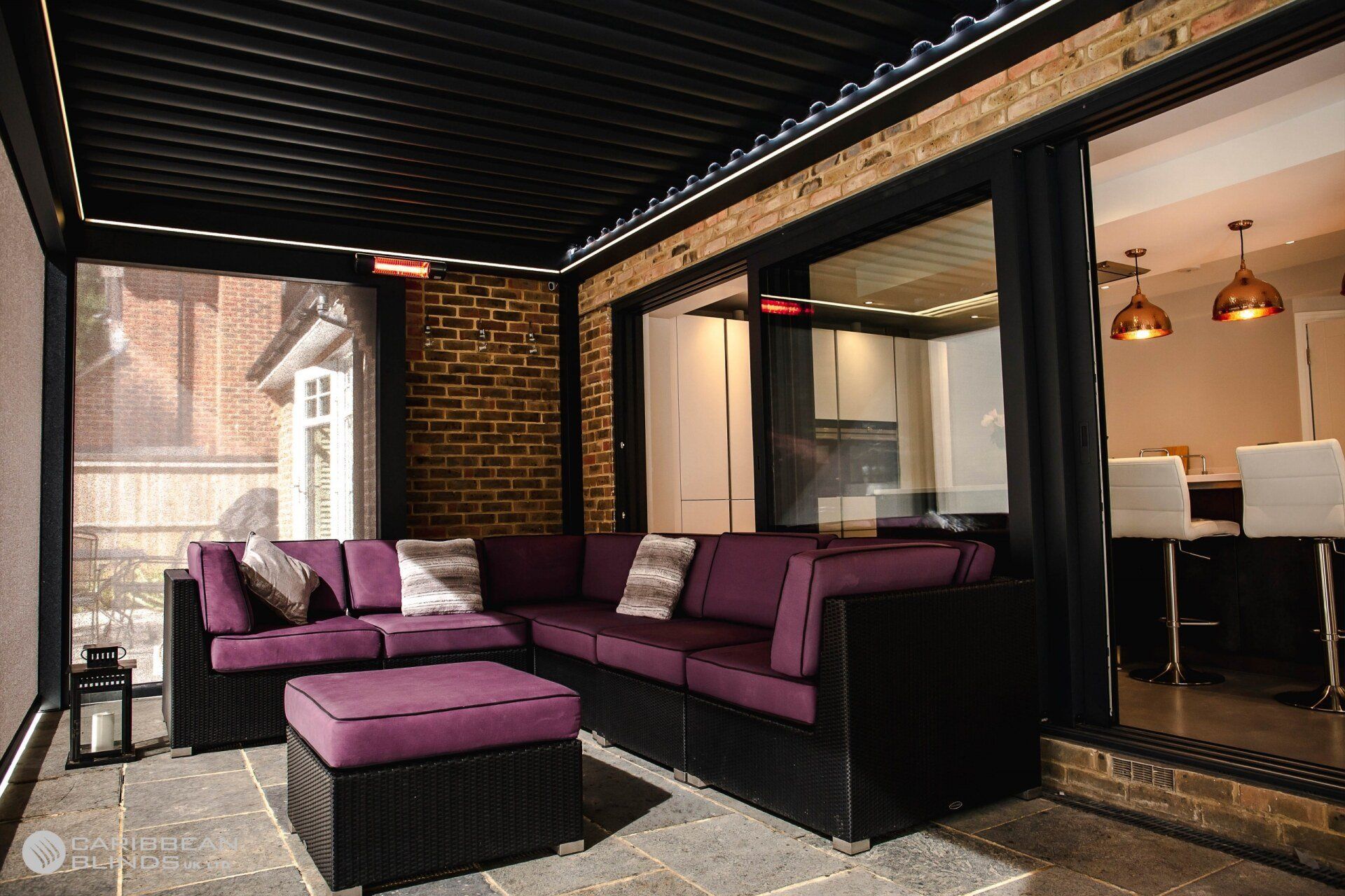 A modern outdoor patio featuring a purple sectional sofa and ottoman under a black pergola against a brick house wall.