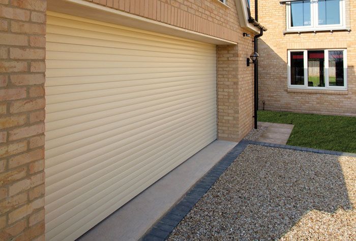 A cream-colored roller garage door set into a brick house next to a gravel driveway and a grassy lawn.