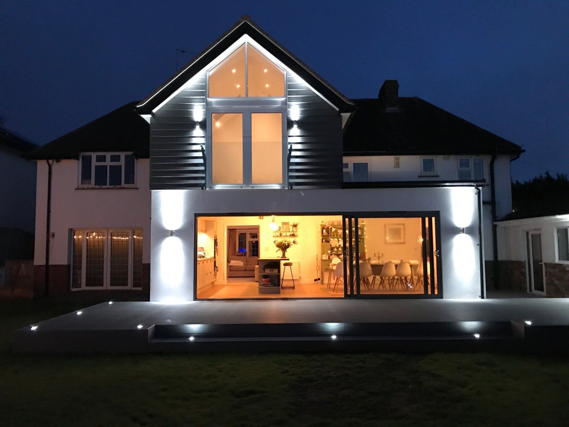 A modern two-story home at night with an illuminated deck, large glass sliding doors, and bright exterior wall lighting.