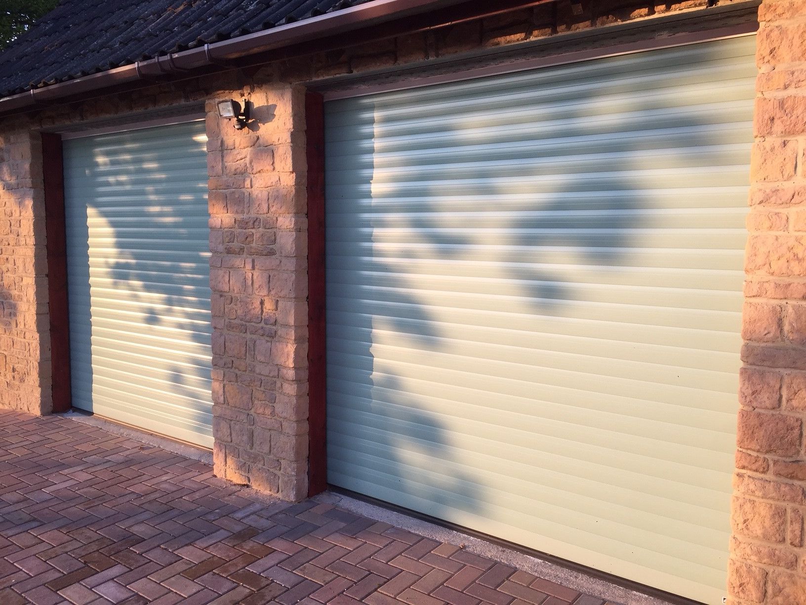 Two light green roller garage doors set in a rustic brick building with tree shadows cast across the surface.