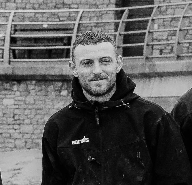 A black-and-white headshot of a person in a hooded work jacket smiling softly, posed in front of a stone wall.