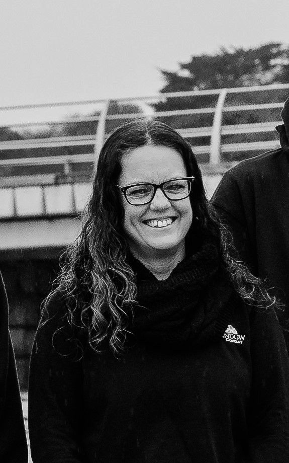 A smiling person with long curly hair and glasses, wearing a dark top, standing outdoors in front of a bridge railing.