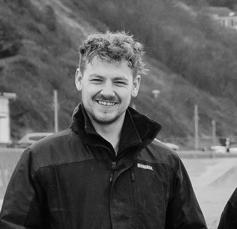A smiling person with curly hair and a beard wearing a zip-up jacket, outdoors in front of a hillside.