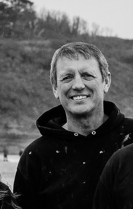 A black-and-white headshot of a smiling person wearing a hooded sweatshirt outdoors with a blurred landscape background.