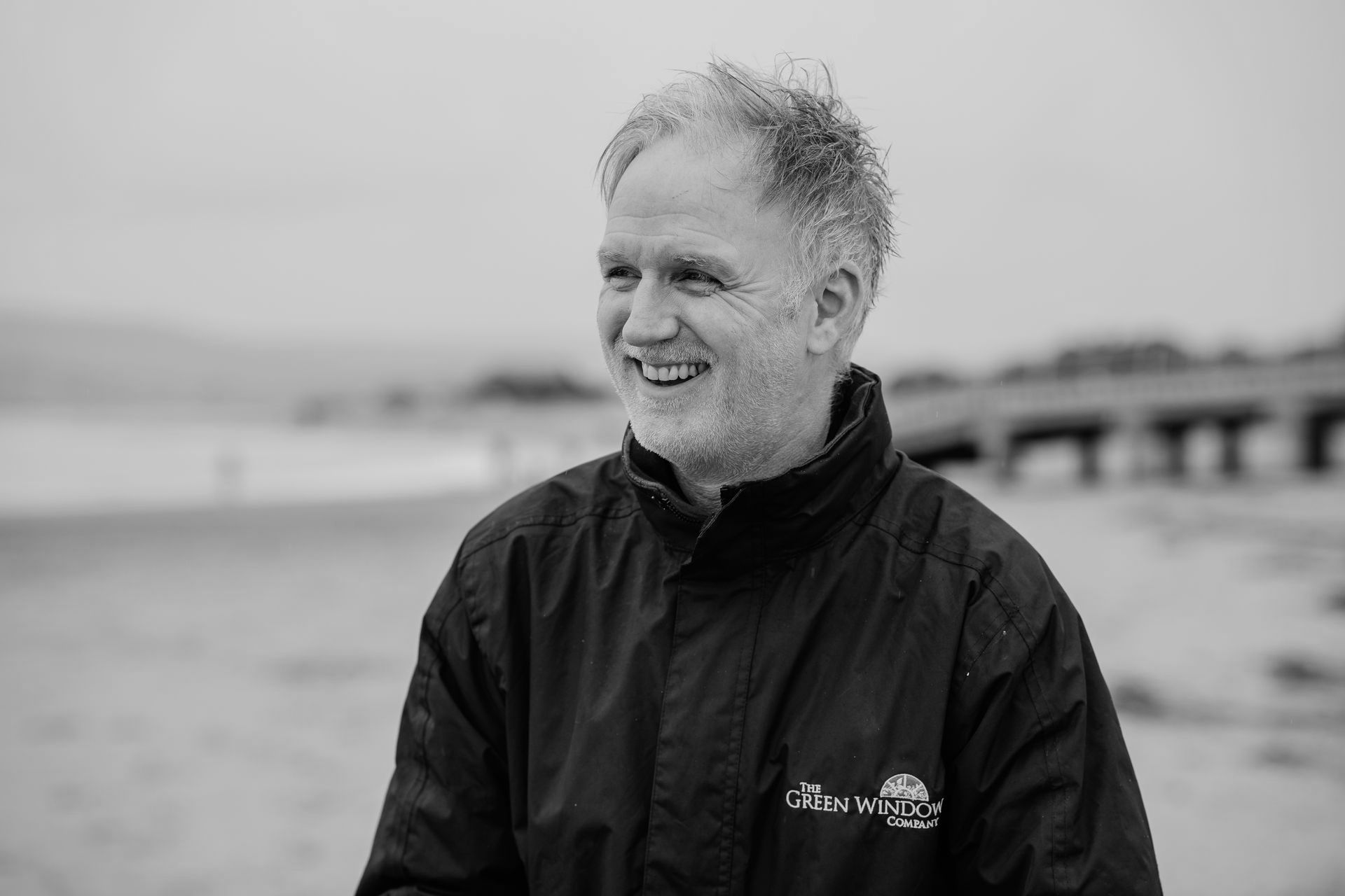 A smiling person in a dark jacket stands on a beach, with a pier visible in the soft-focus background.