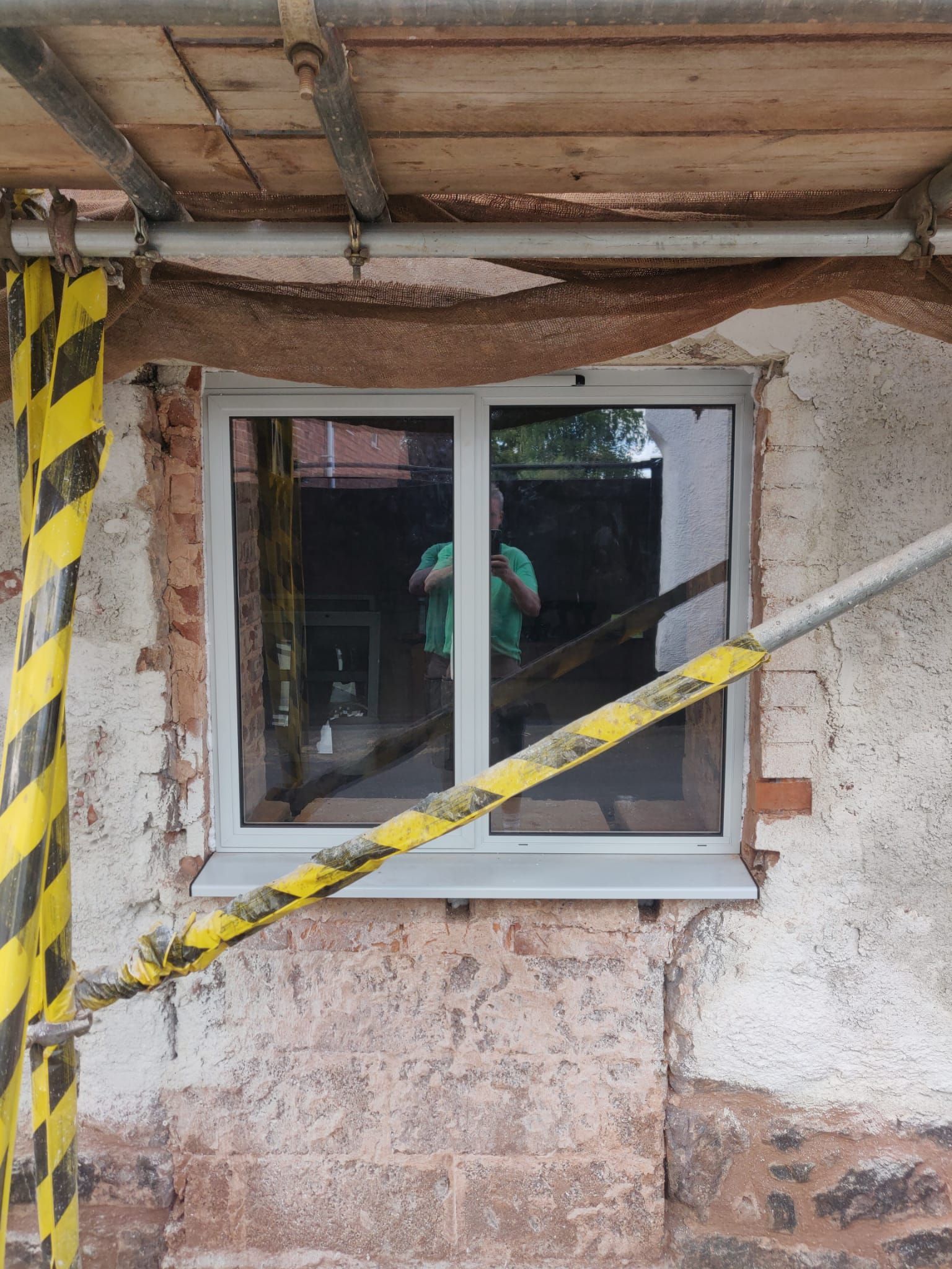 A white window frame is installed in a rough, unplastered wall surrounded by yellow and black hazard caution tape.