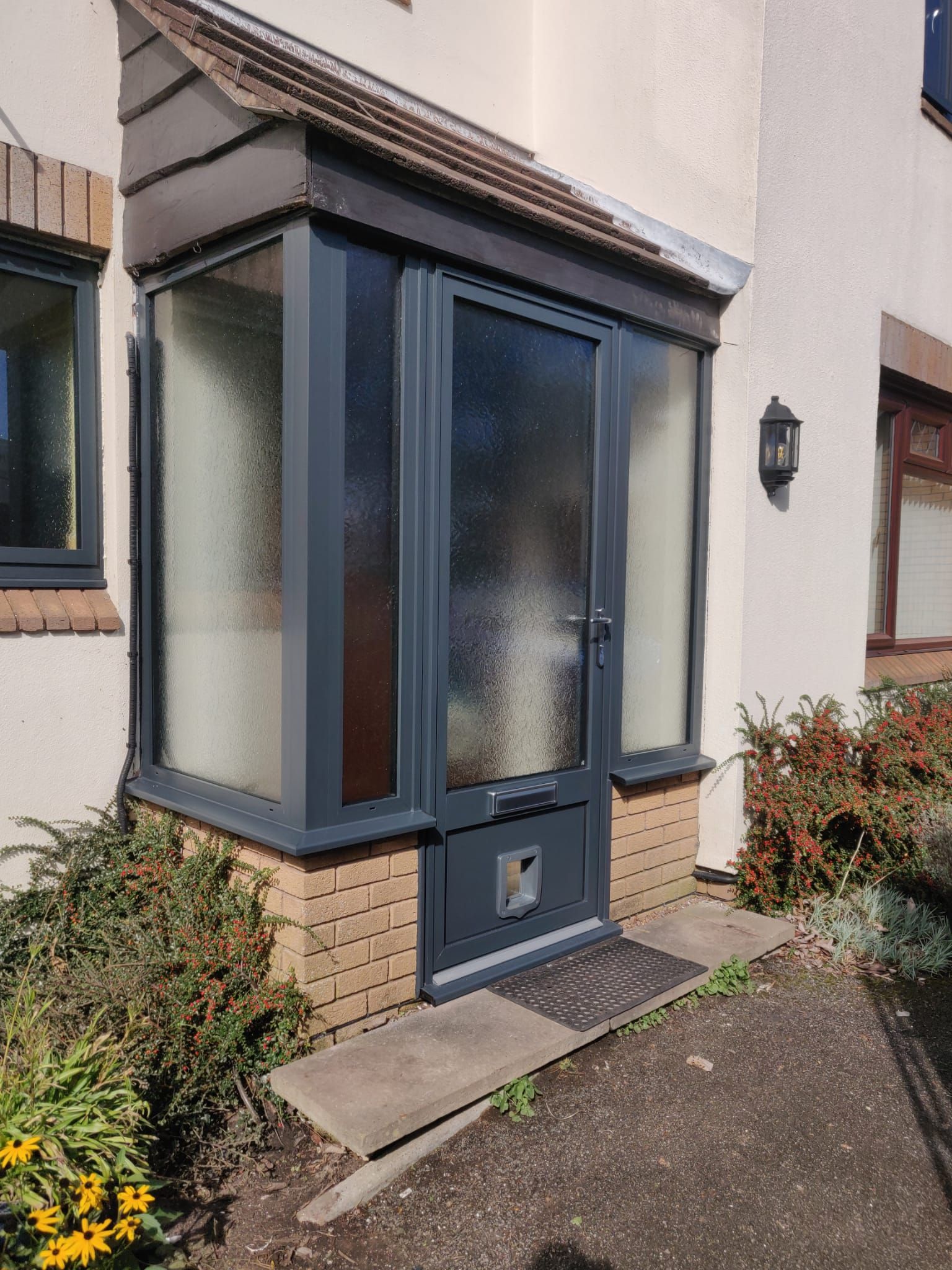 A dark grey porch extension with frosted glass panels and a front door, built onto a house with a brick base.