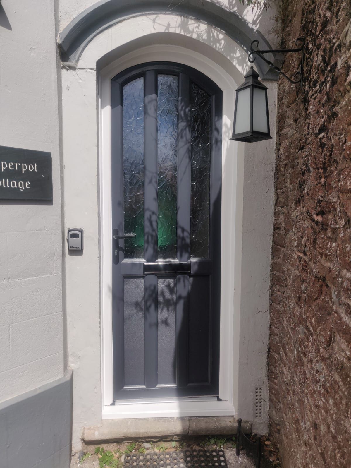 A dark grey, arched front door with decorative glass panes in a white frame, next to a black lantern and brick wall.