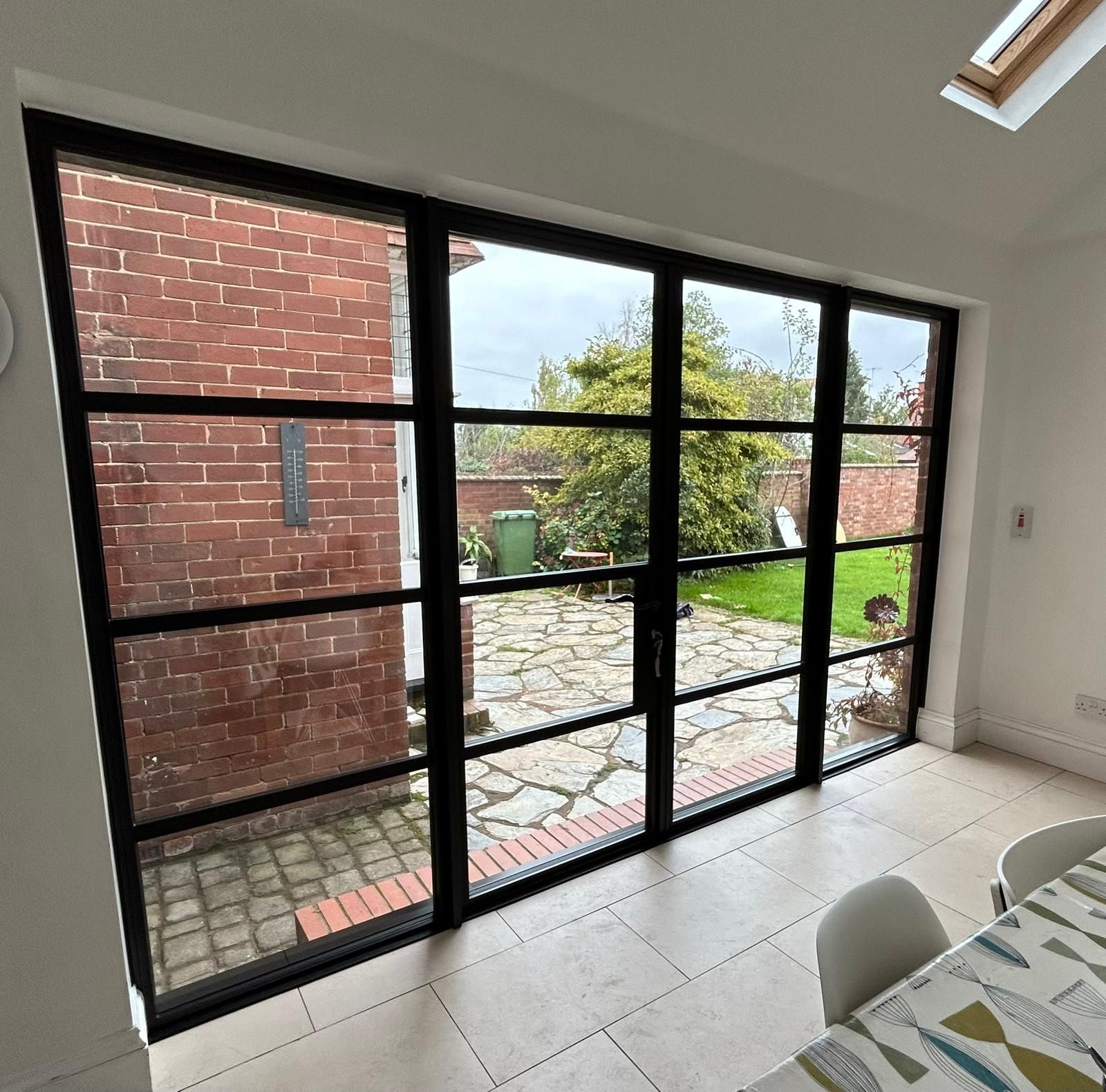 Large black-framed glass folding doors open onto a stone patio and garden from a brightly lit, modern dining room.