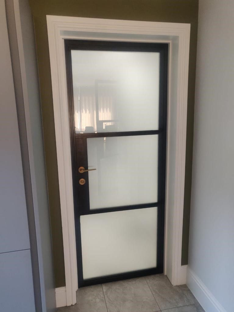 A black-framed door with three frosted glass panels installed in a white door frame against a dark olive wall.