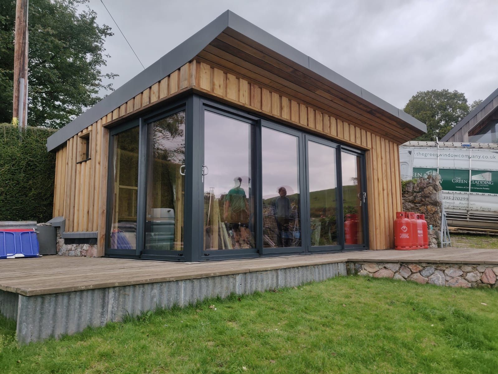 A modern garden room with vertical wood siding and large sliding glass doors, situated on a wooden deck in a grassy yard.