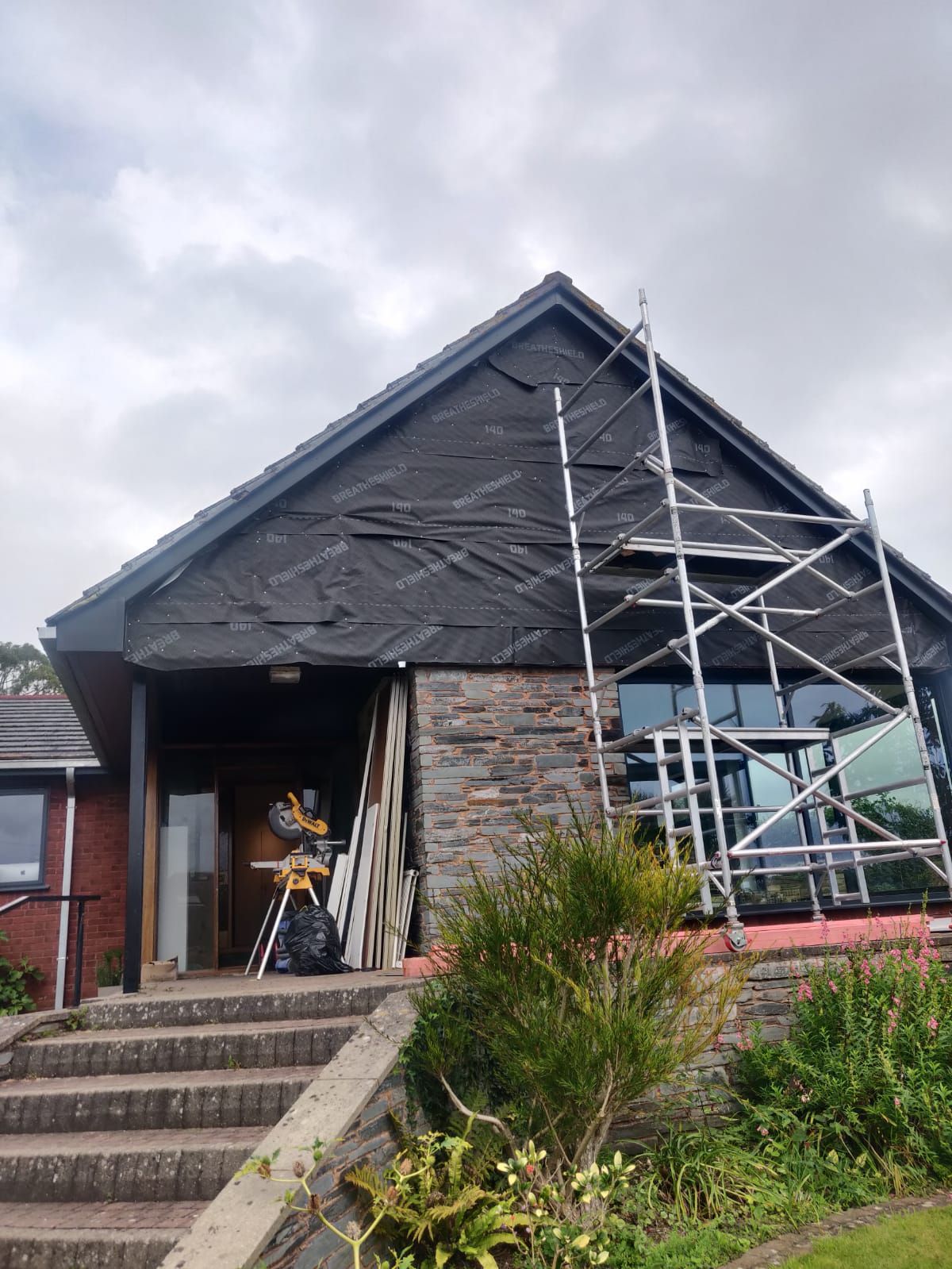 Scaffolding stands before a house with a stone lower wall and a black weather-resistant membrane covering the gable end.