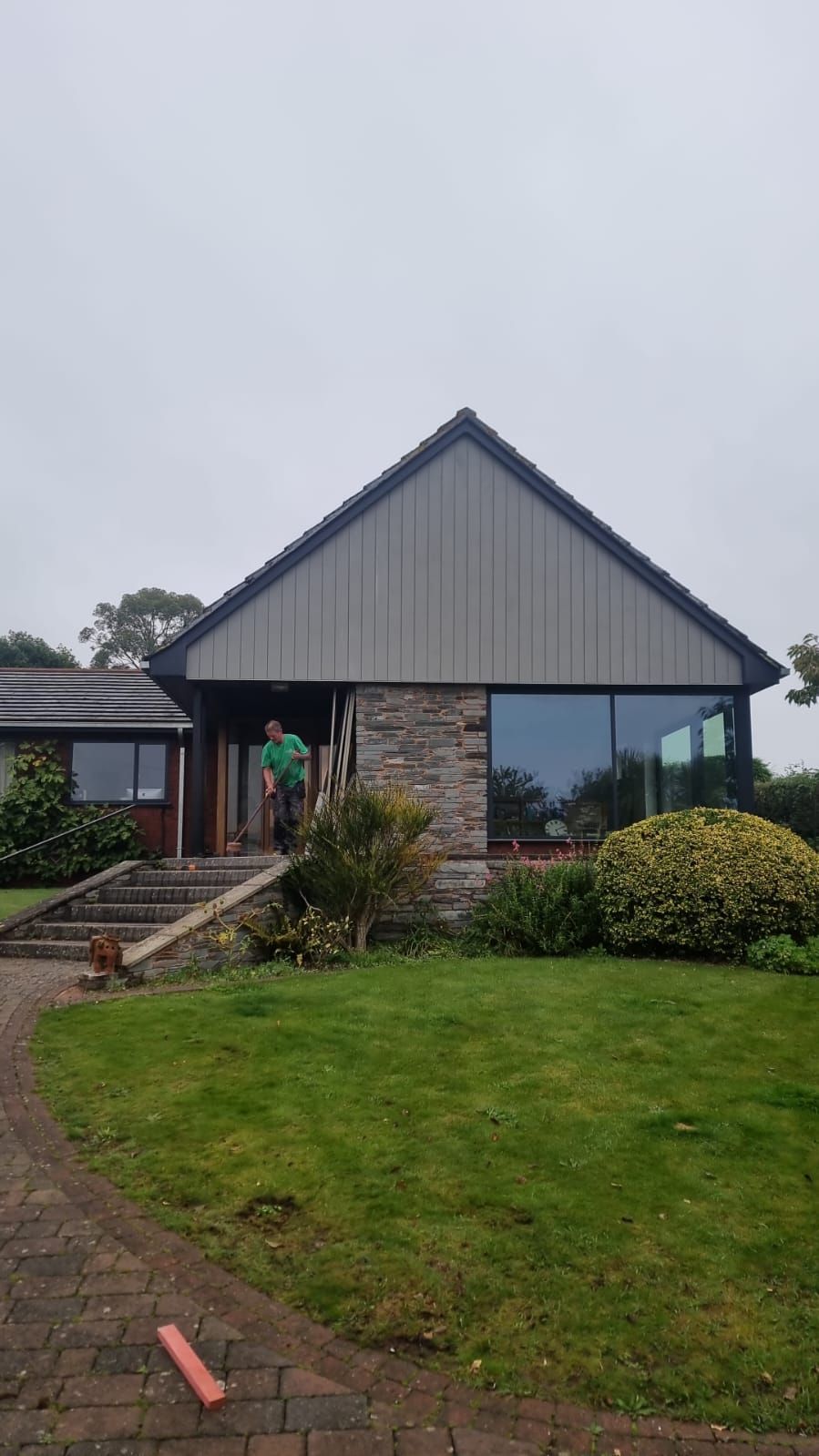 A person in a green shirt stands by the stone entryway of a modern house with gray vertical siding and a large window.