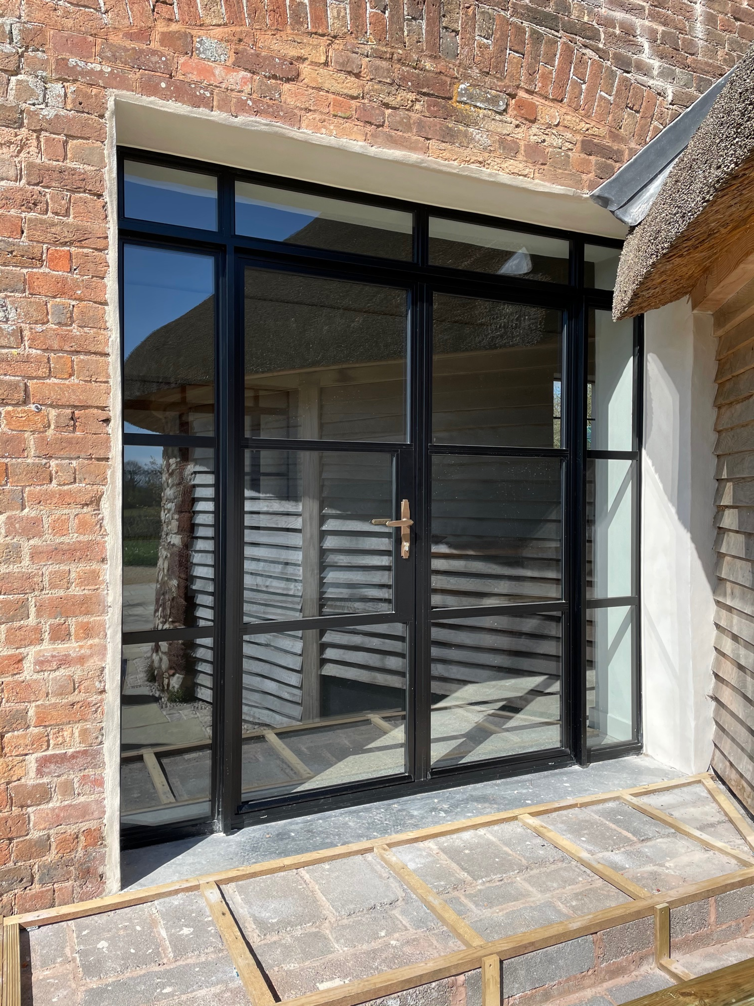 A black-framed glass door and side panels set into a brick wall next to a wooden building with a thatched roof.